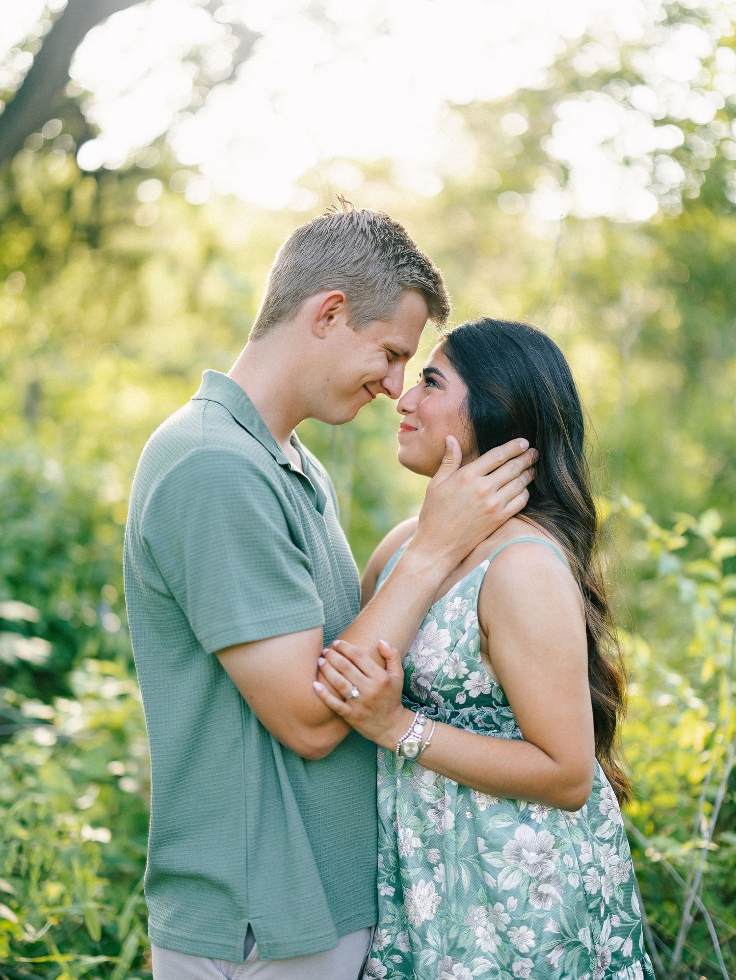 Couple sharing a romantic moment outdoors, touching foreheads in a lush green garden during golden hour.