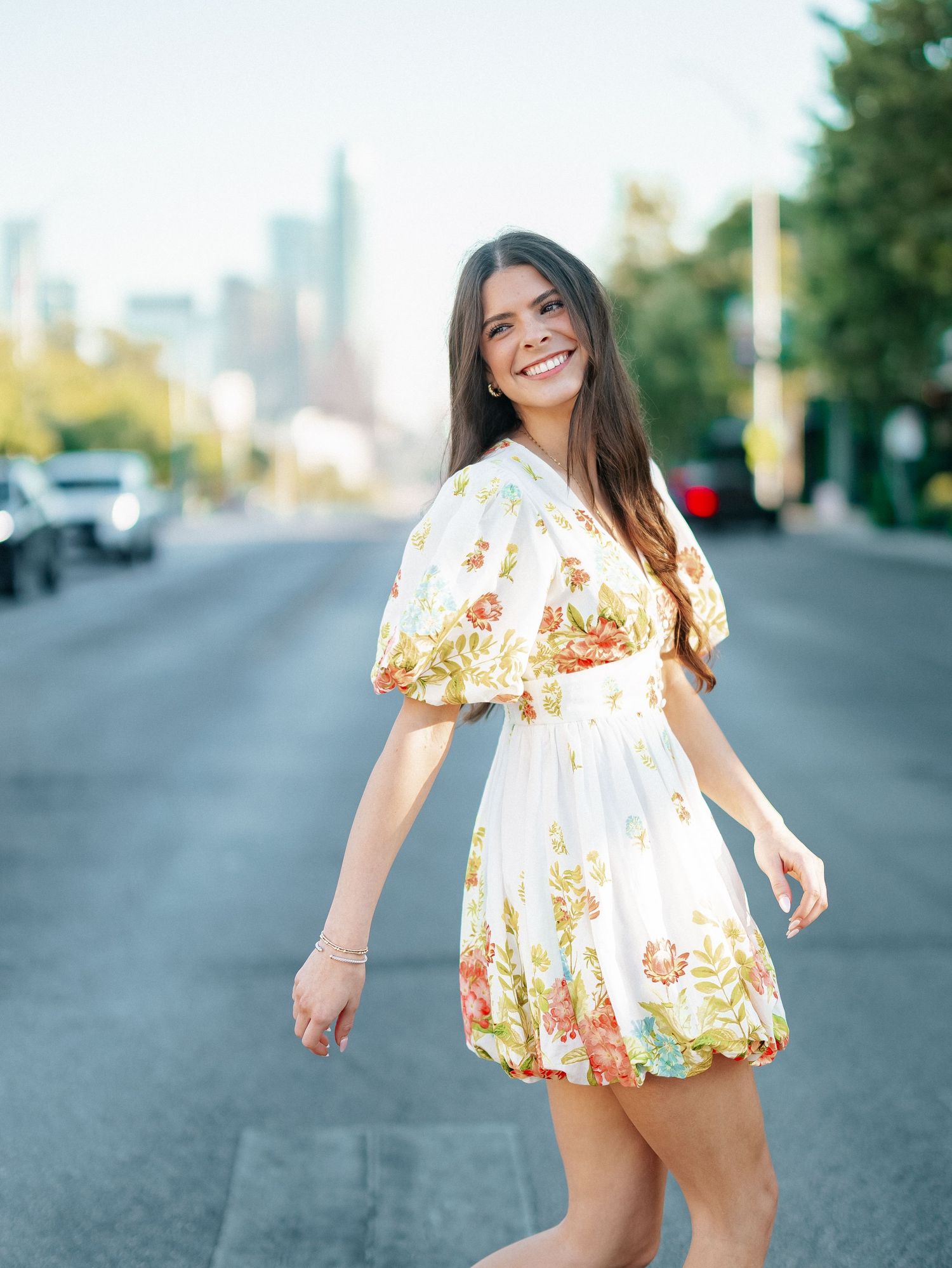 Brunette woman in white floral dress smiling while walking on city street with skyline backdrop.