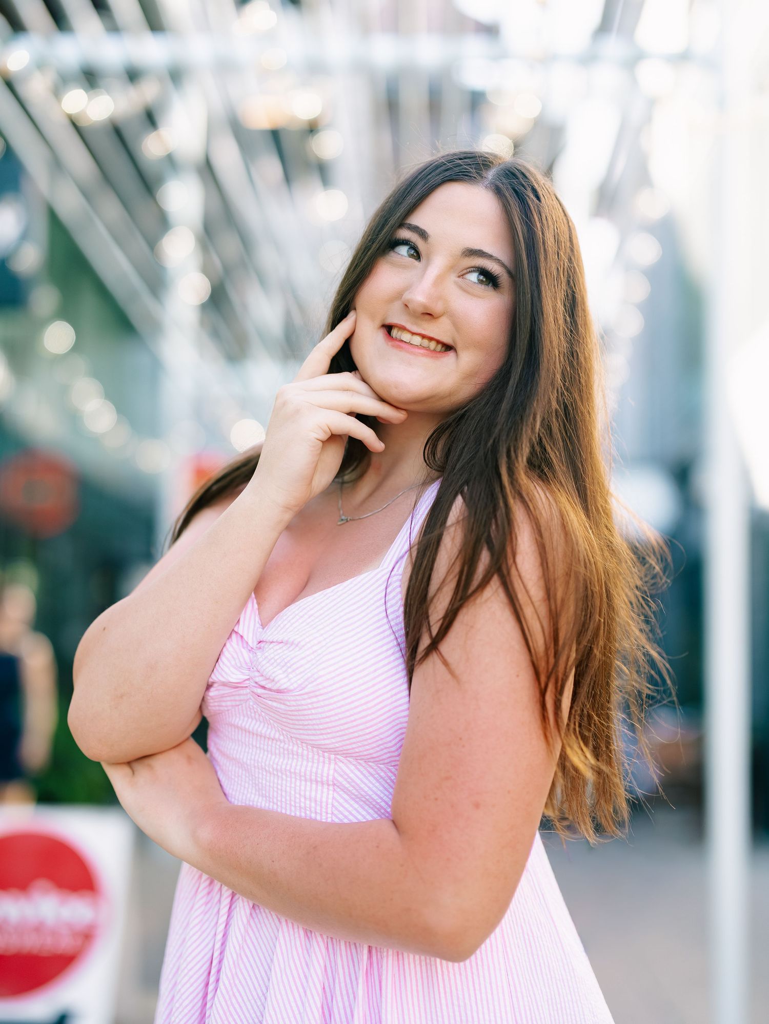 Brunette woman in pink striped dress smiling and touching face in bright outdoor setting.