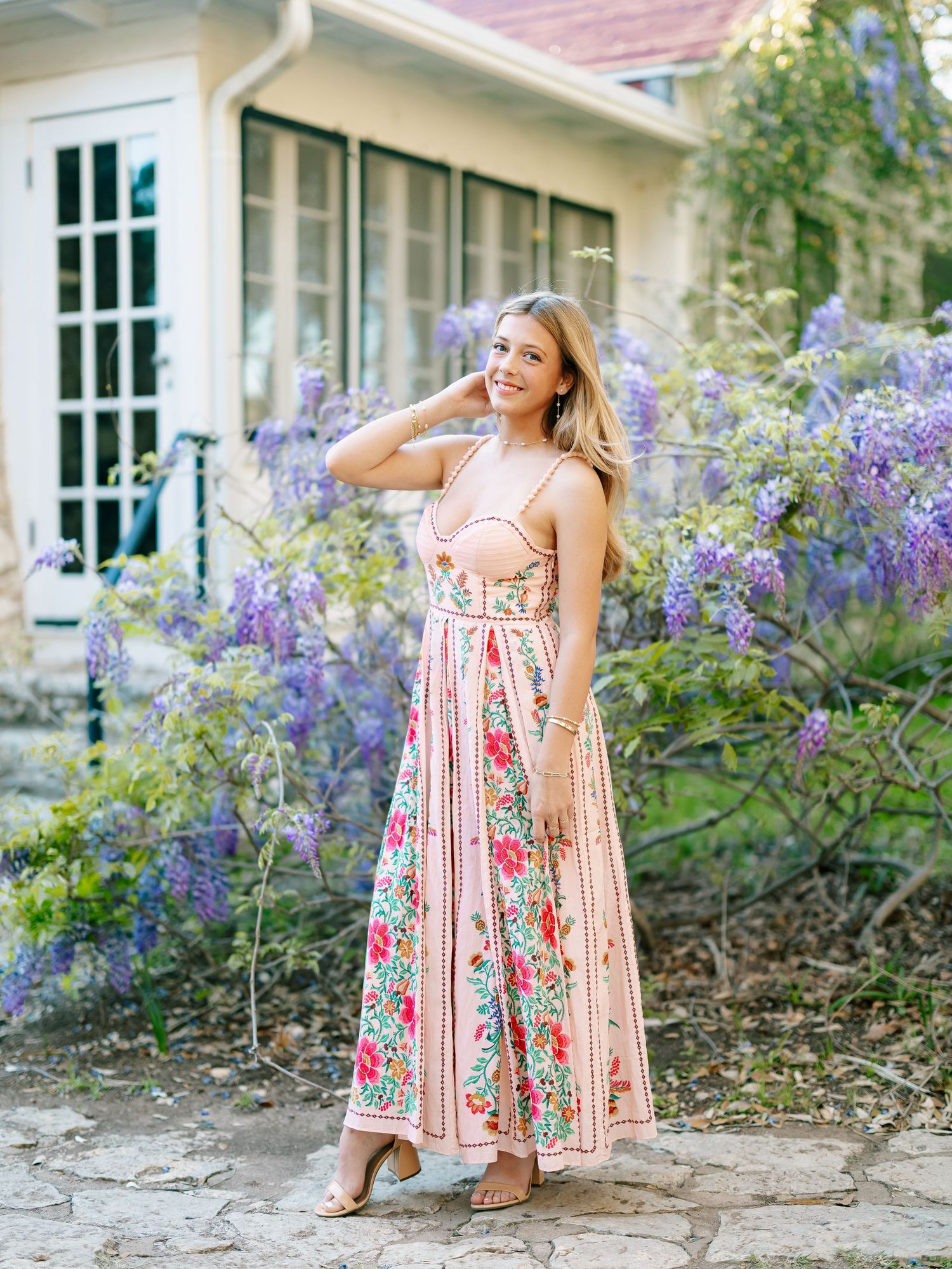 Smiling blonde woman in pink floral maxi dress standing before purple wisteria blooms.