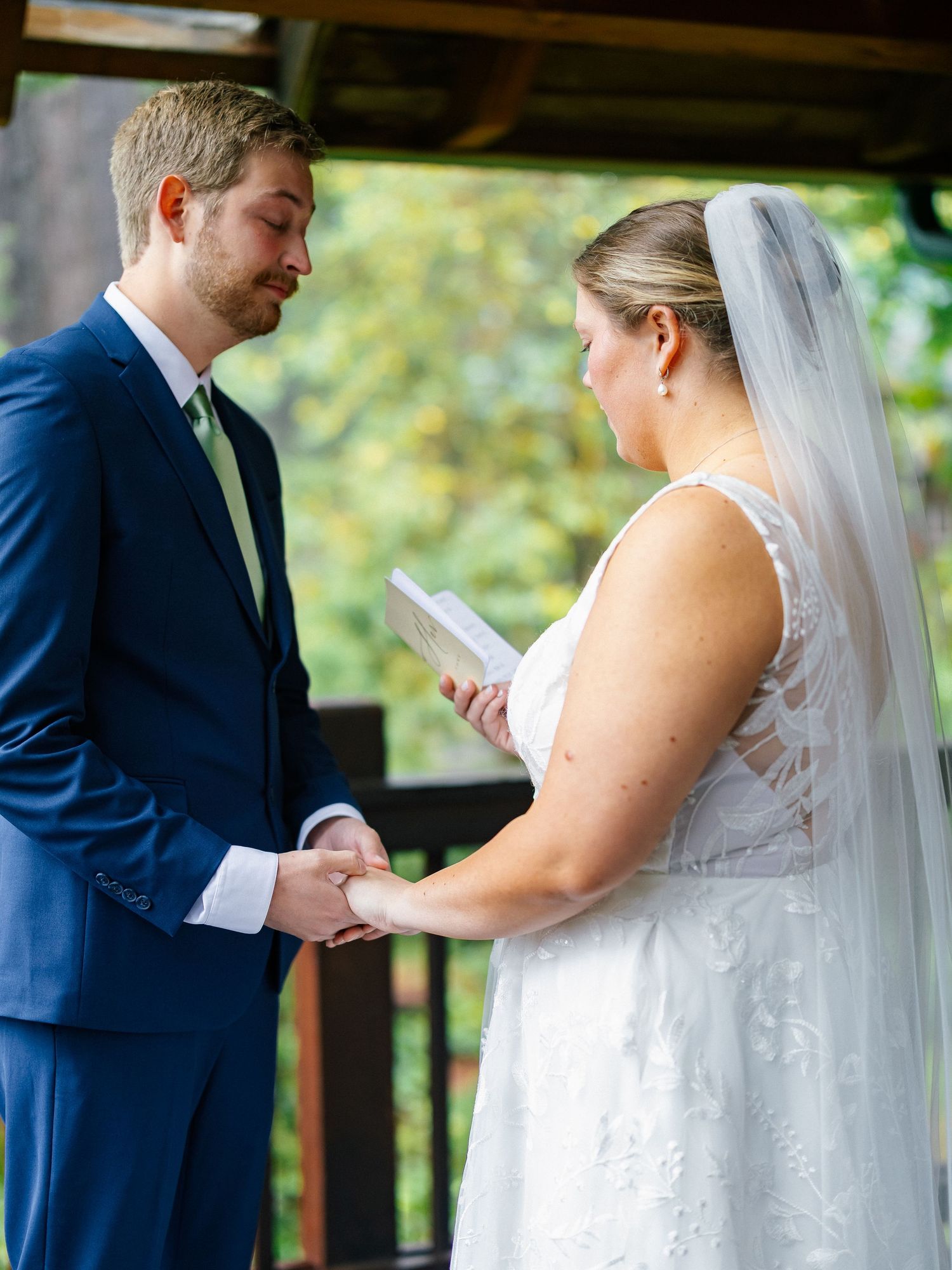 Groom holds bride's hands as she reads heartfelt vows during an outdoor woodland wedding ceremony.