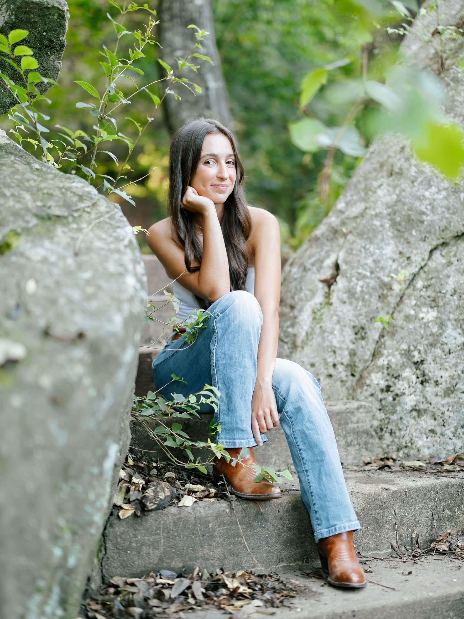 Brunette woman in gray tank and blue jeans sitting on stone steps surrounded by lush greenery.