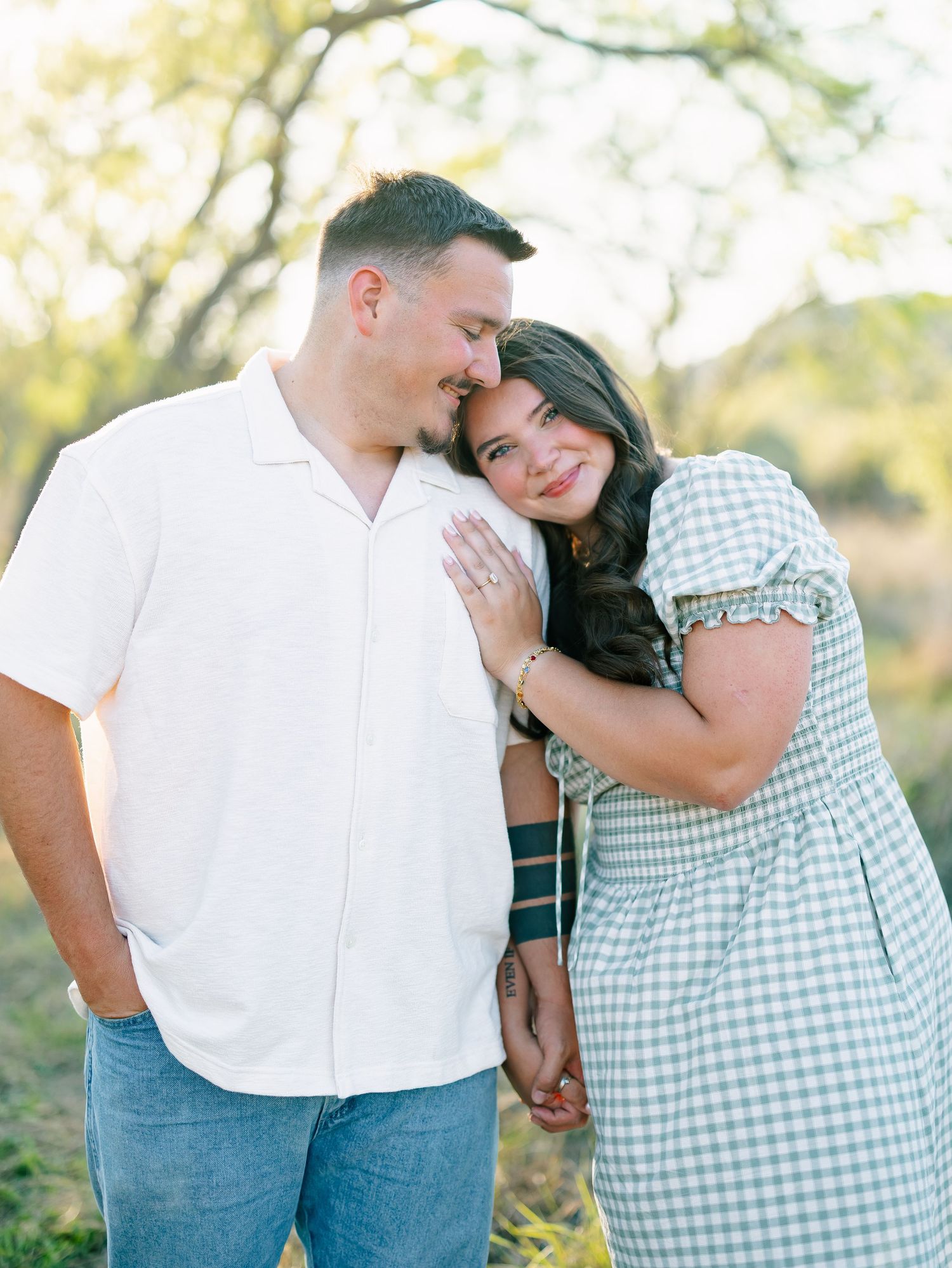 A couple shares a tender moment in a sunny field, woman in plaid dress leaning on mans shoulder smiling.