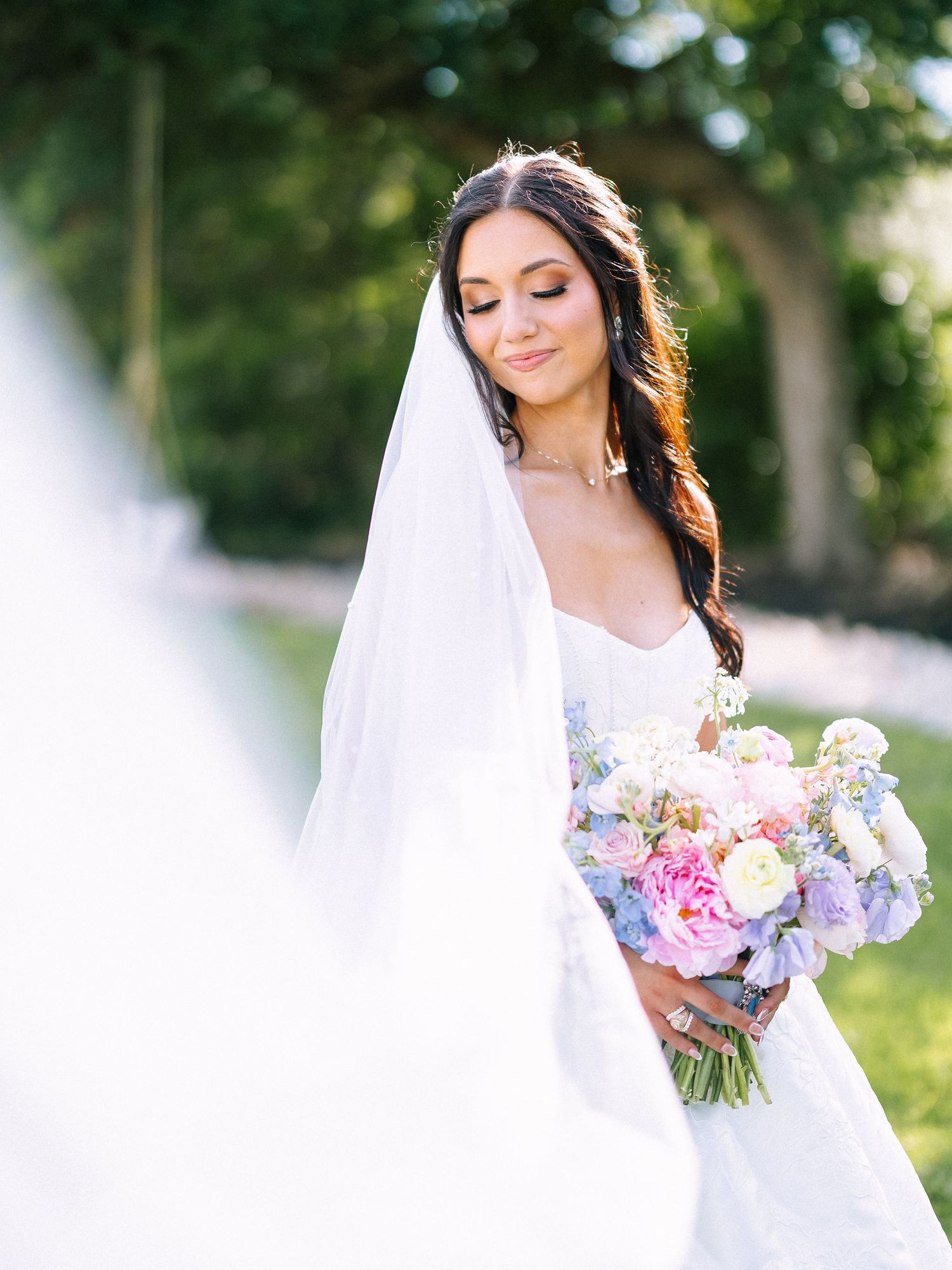Bride with dark hair wearing veil holds colorful pink and blue bouquet outdoors in soft sunlight.