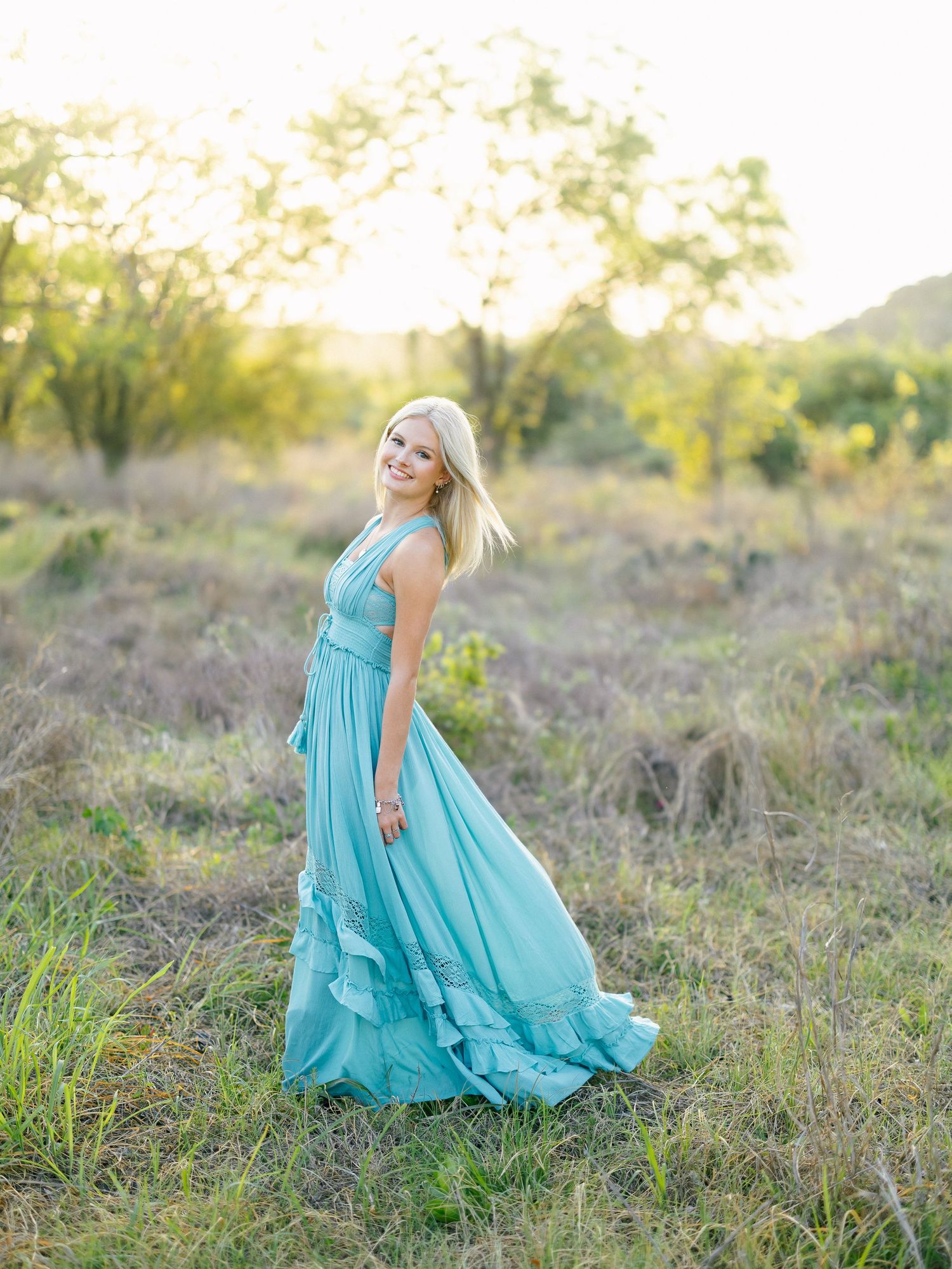 Blonde woman twirling in a flowing turquoise gown in a sunlit meadow at golden hour.