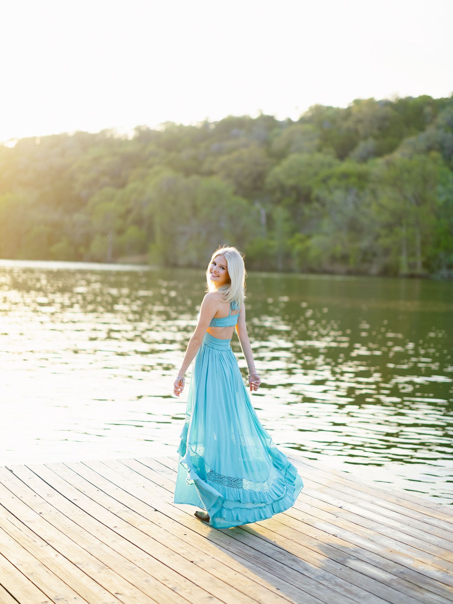 A woman in a flowing turquoise two-piece gown poses on a wooden dock by a sunlit lake.