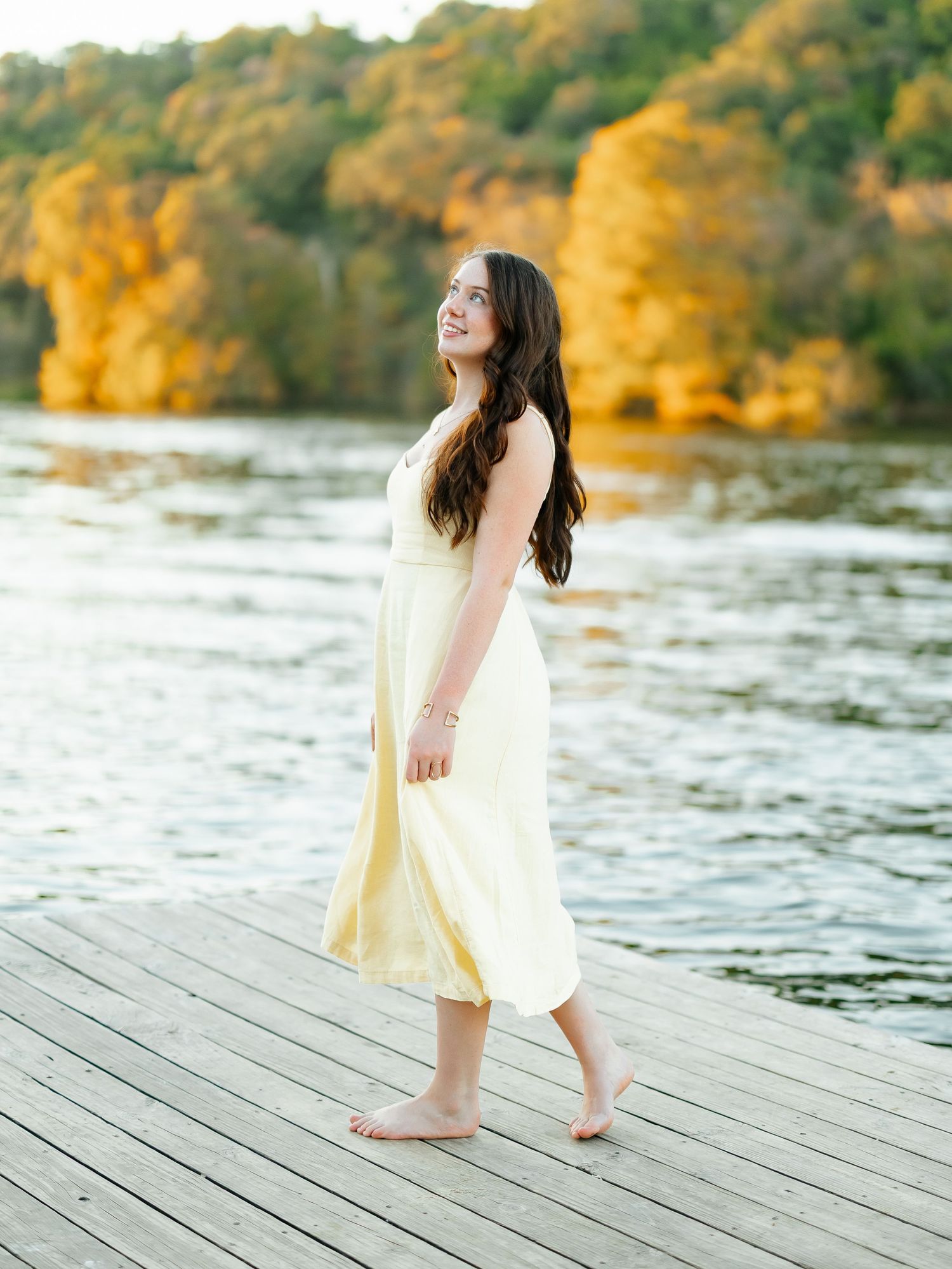 Young woman in yellow dress standing barefoot on wooden dock by autumn lake with colorful foliage.