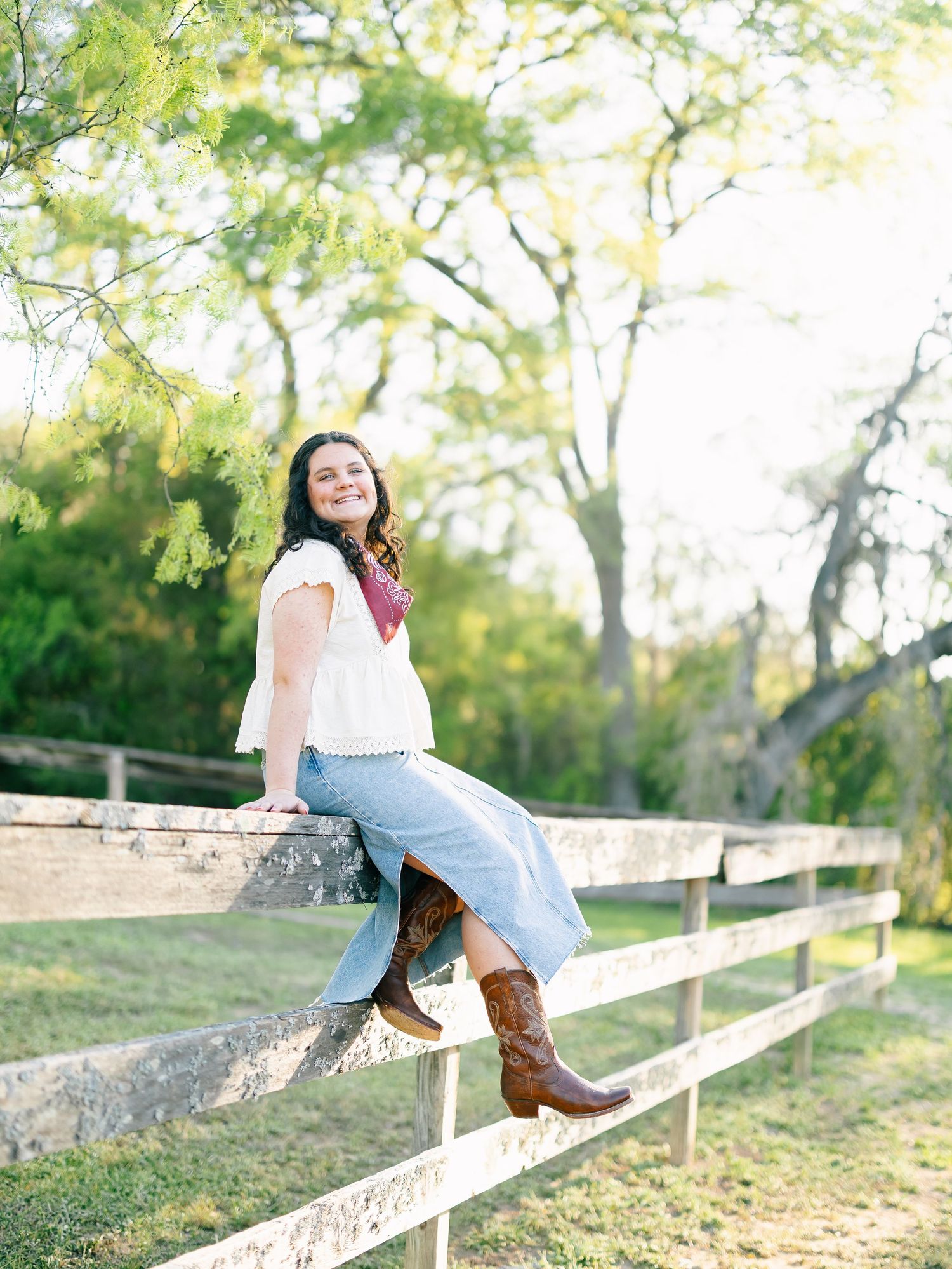 Young woman in white top, jeans, and cowboy boots sitting on a wooden fence outdoors with green trees behind her.
