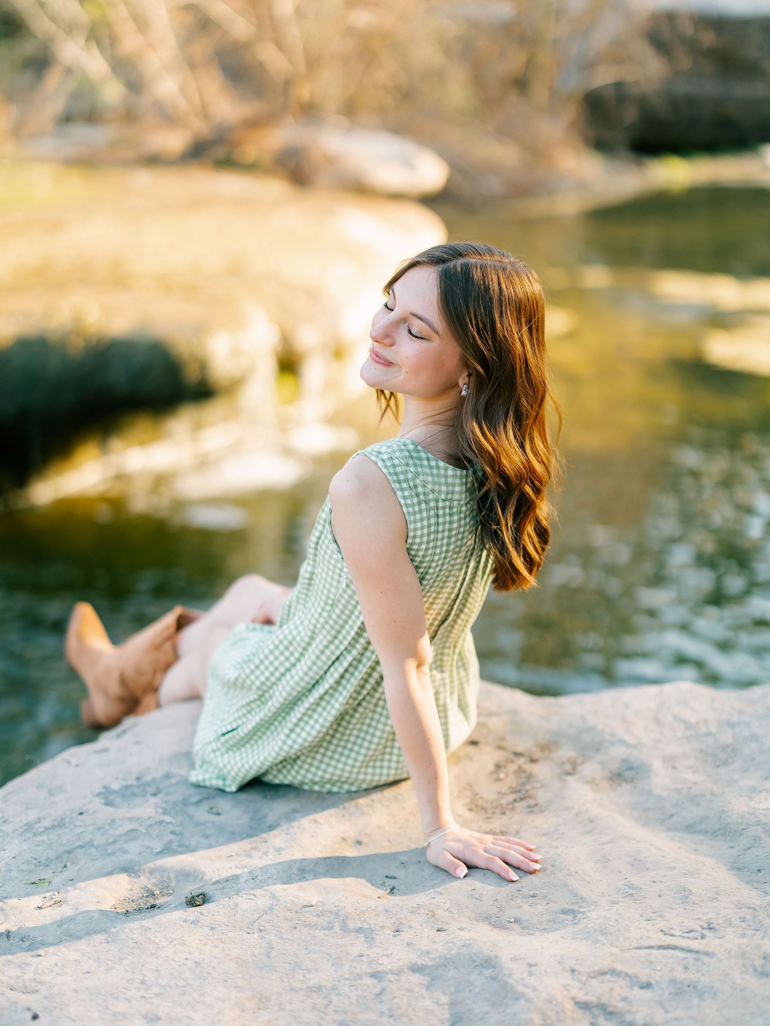 Young woman in green dress and cowboy boots sitting on rocks by a serene creek in golden hour light.