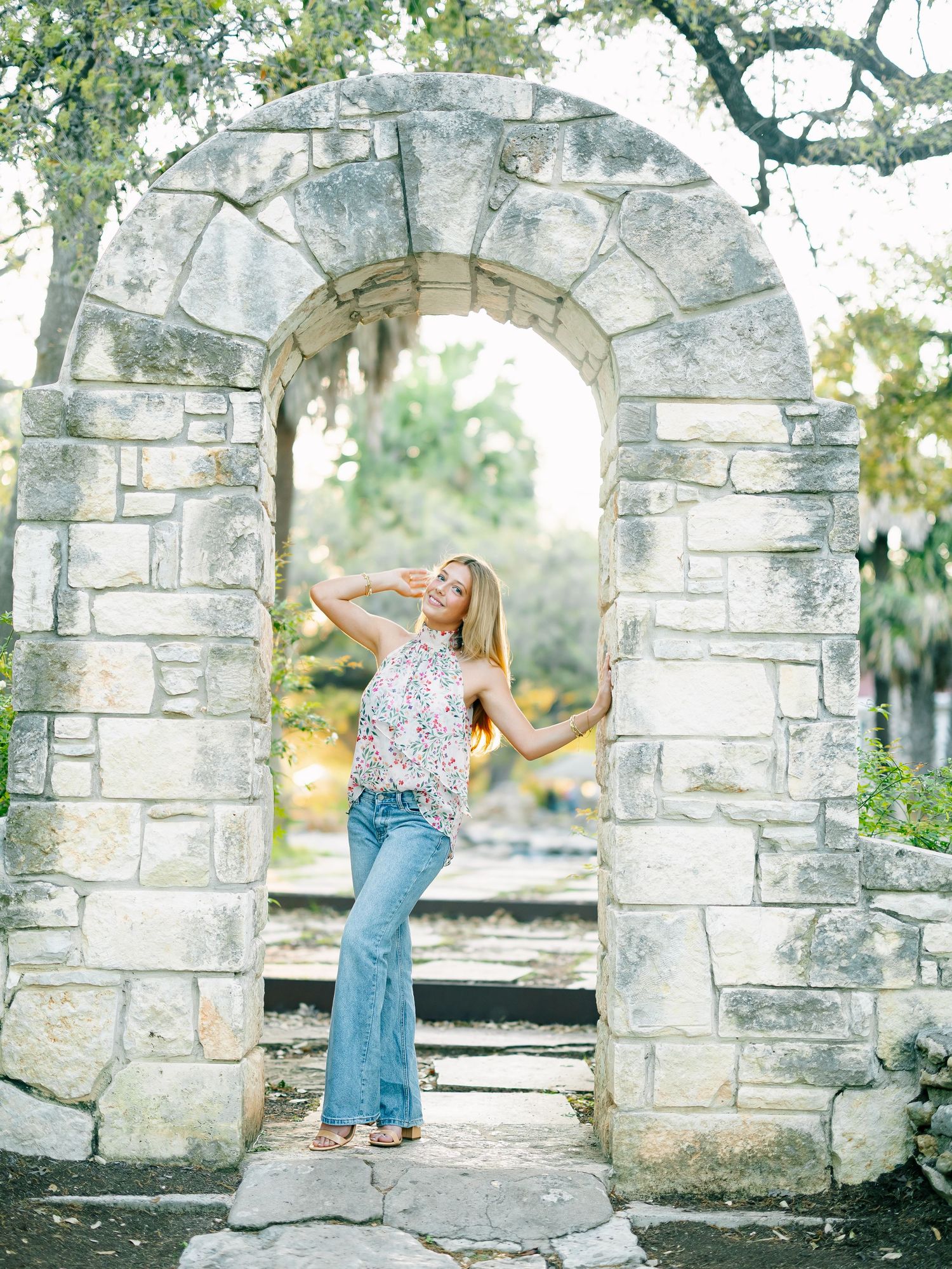 Young woman in floral top and flared jeans posing under a rustic stone arch in a lush outdoor garden setting.