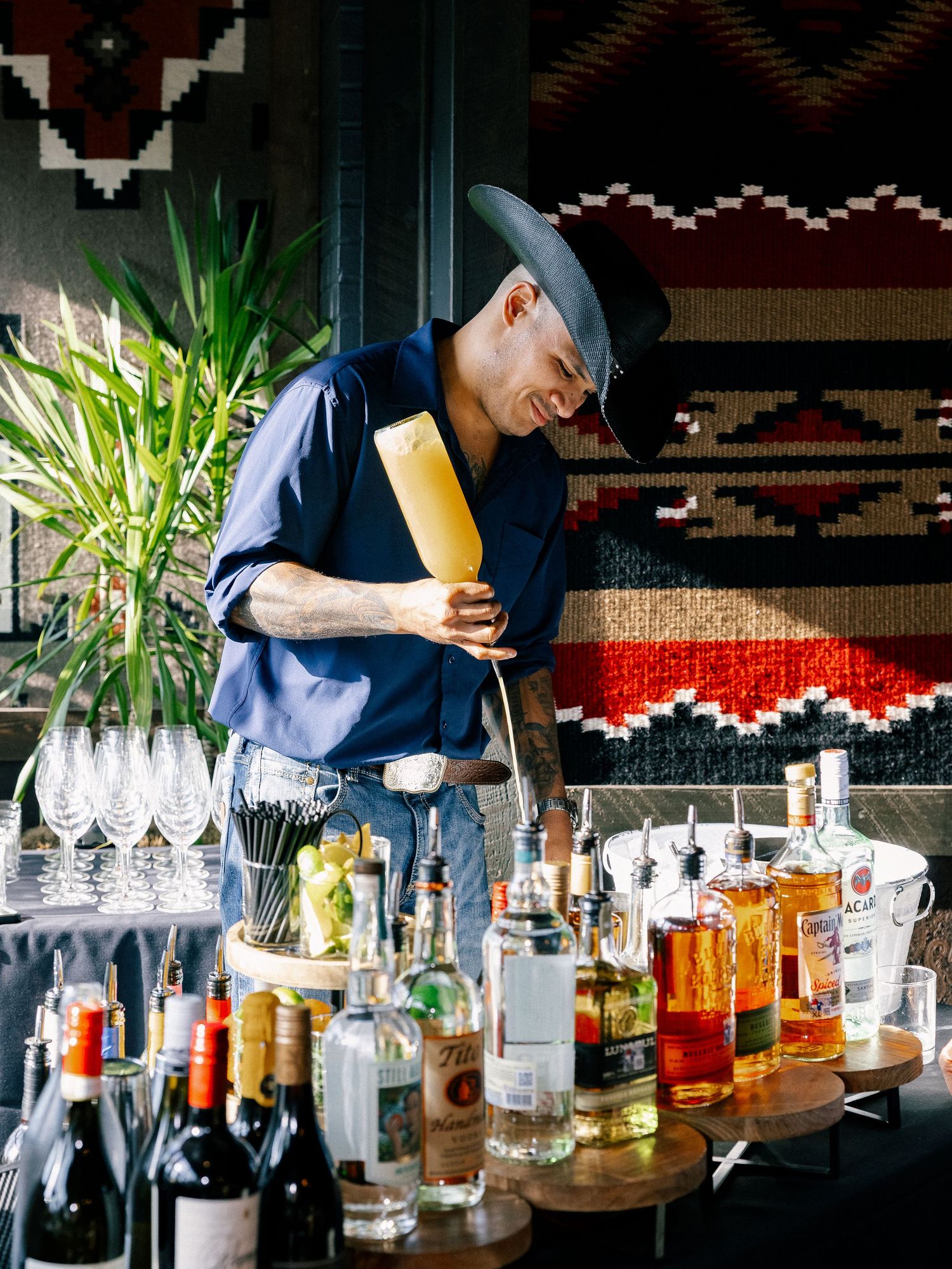 Bartender in gray hat pouring a cocktail surrounded by bottles at a stylish bar with colorful tapestry backdrop.