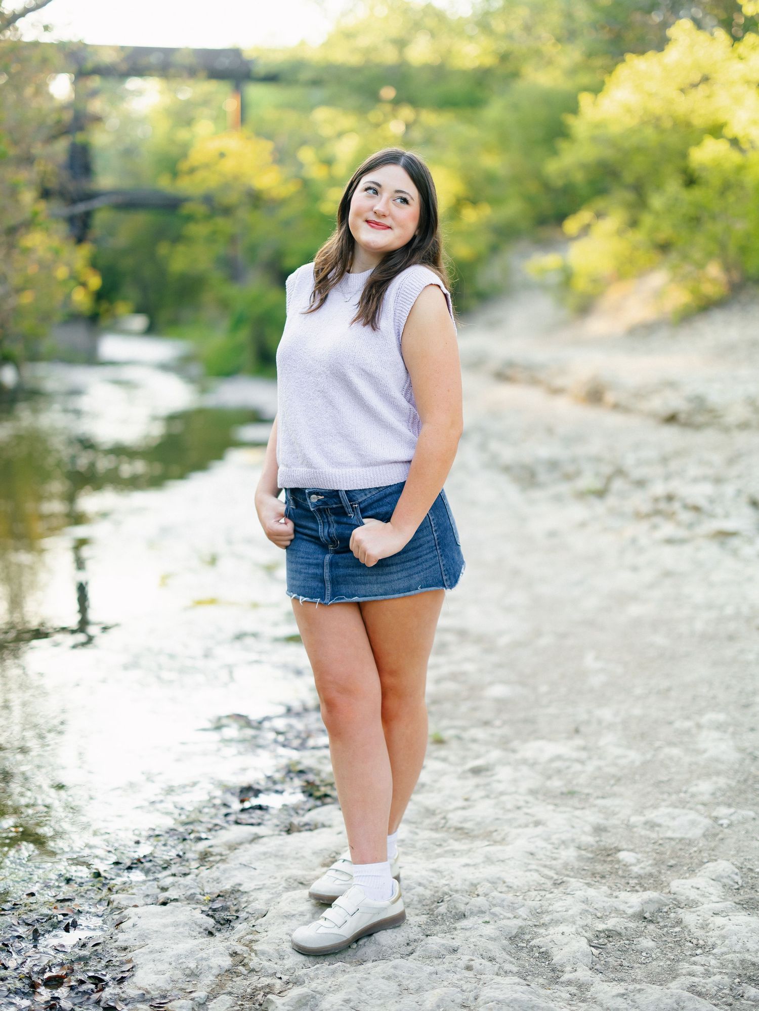 Smiling young woman in white top and denim skirt standing by a scenic creek.