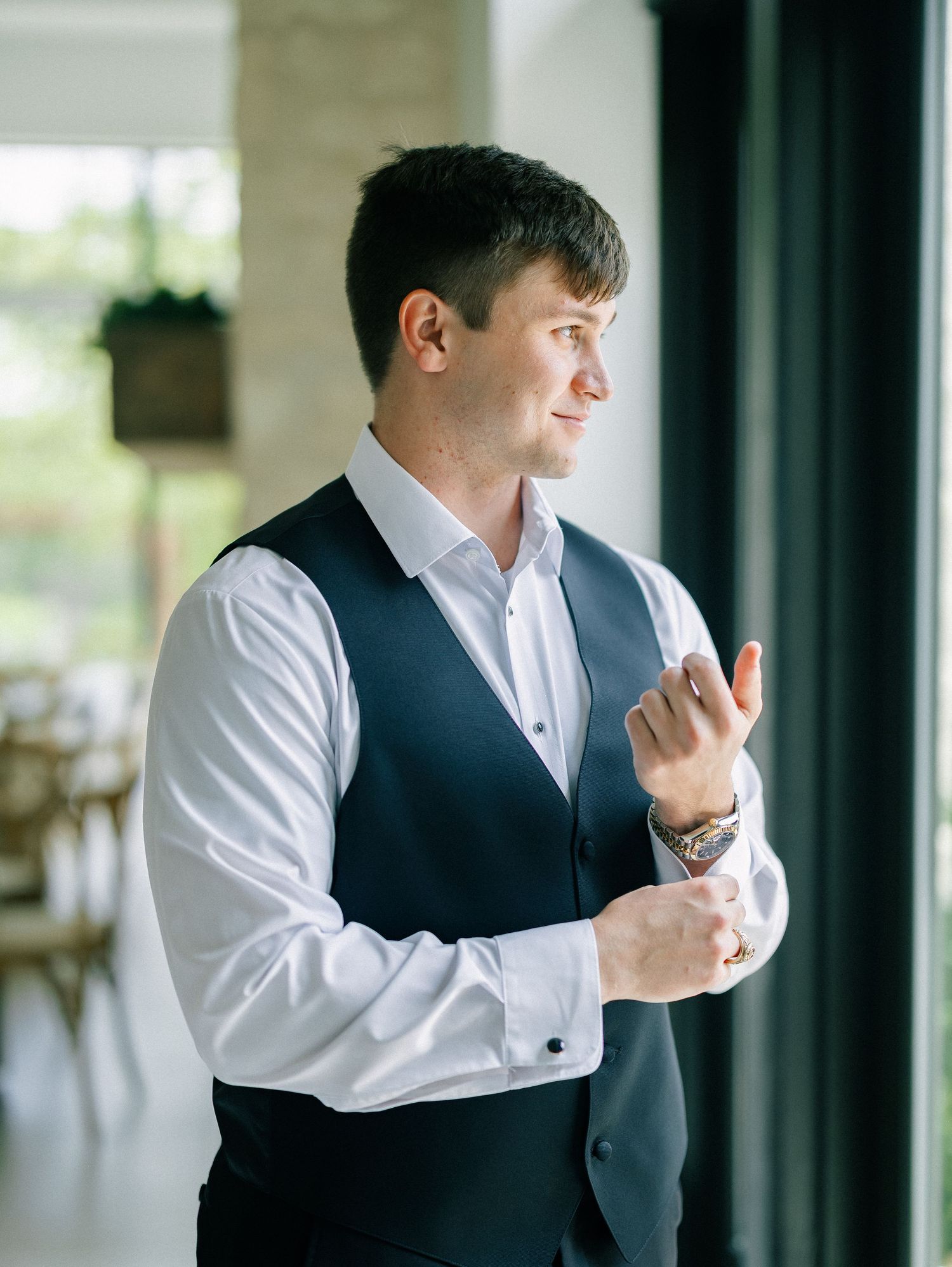 A well-dressed man in a white shirt and navy vest adjusting his cufflinks near a bright window.
