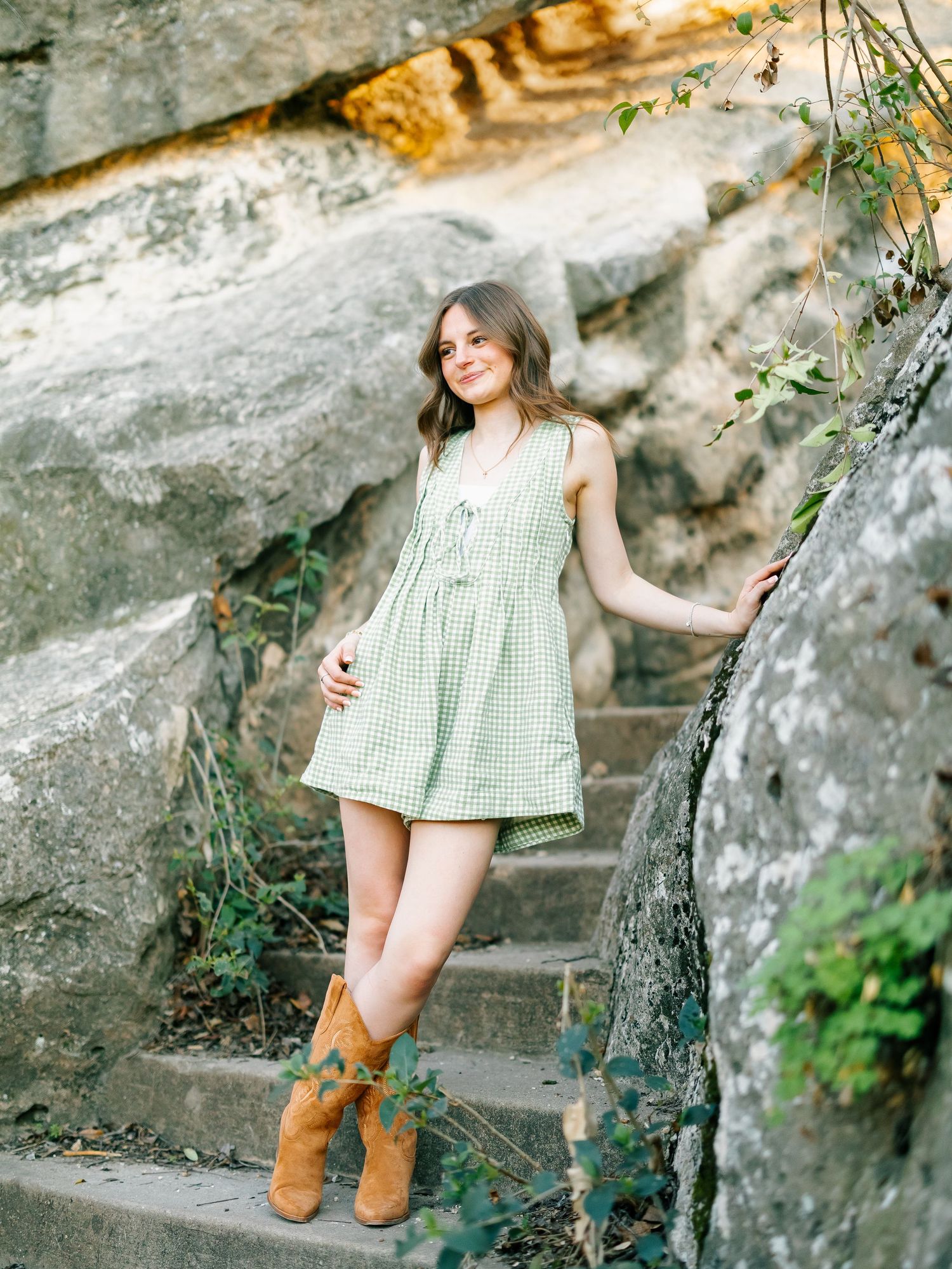 Young woman in green gingham dress and brown cowboy boots posing on stone steps surrounded by rocks.