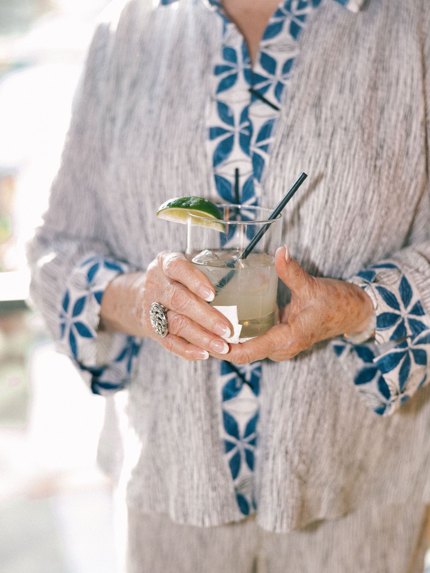 Elderly woman holding a cocktail glass with lime garnish and metal straw, wearing a blue floral patterned jacket.