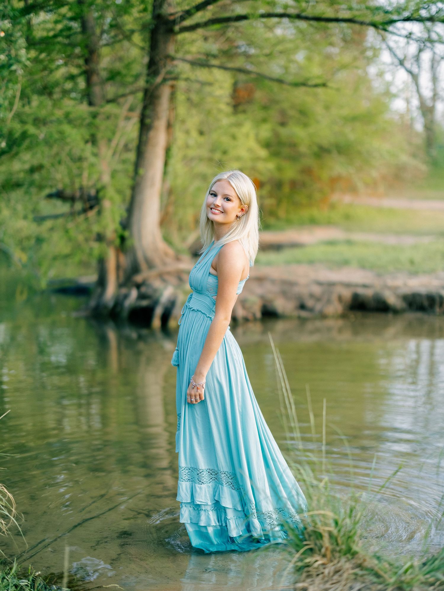 Blonde woman in long turquoise dress standing in shallow creek surrounded by trees.