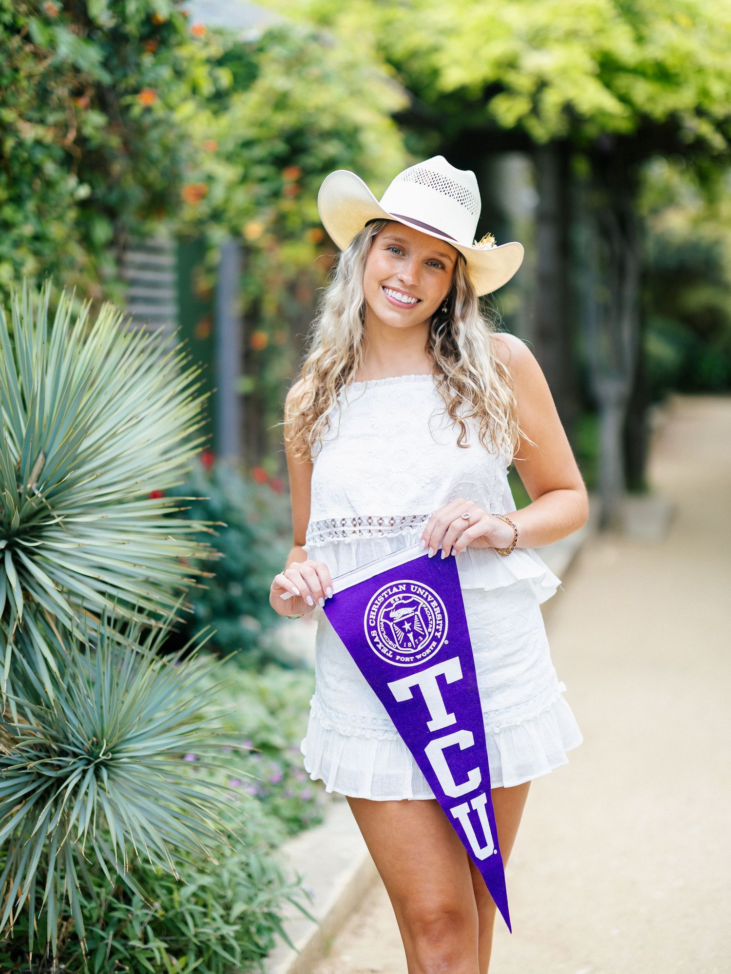 Smiling blonde woman in white dress and cowboy hat holding a purple TCU pennant flag outdoors.