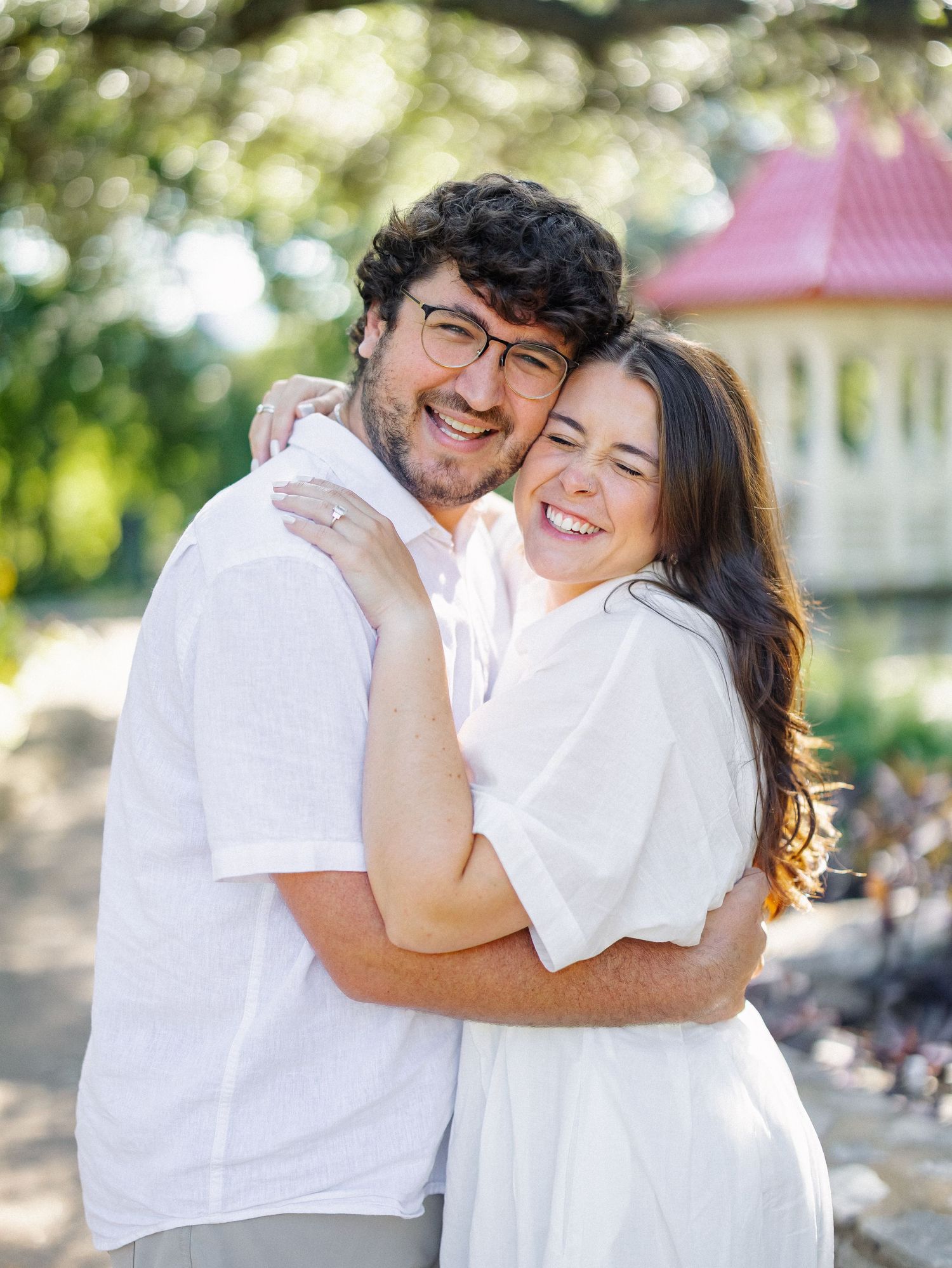 A smiling couple in white outfits embracing outdoors near a pink-roofed building surrounded by lush green trees.
