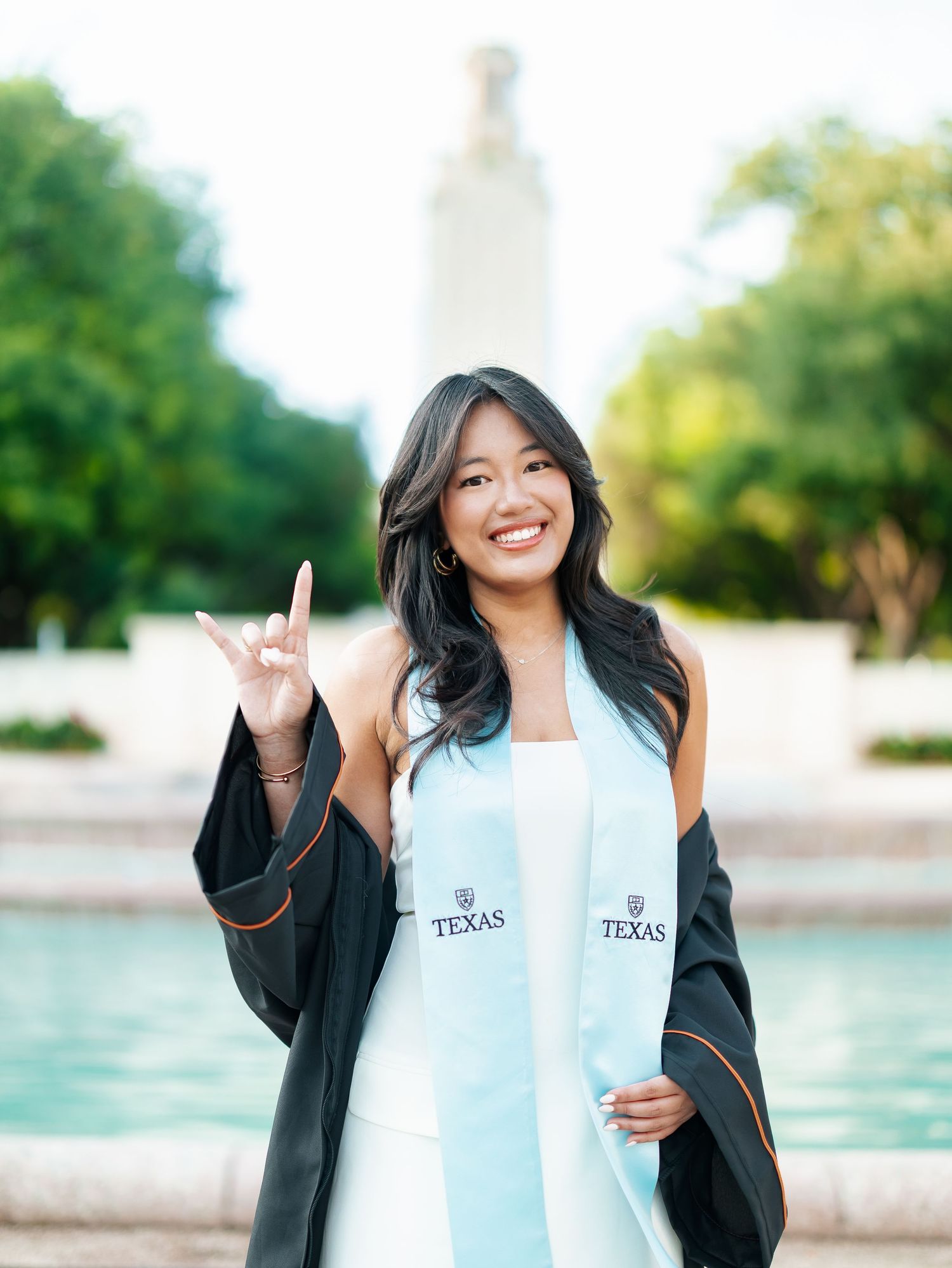 Smiling graduate in cap and gown with Texas stole poses at University of Texas tower fountain.