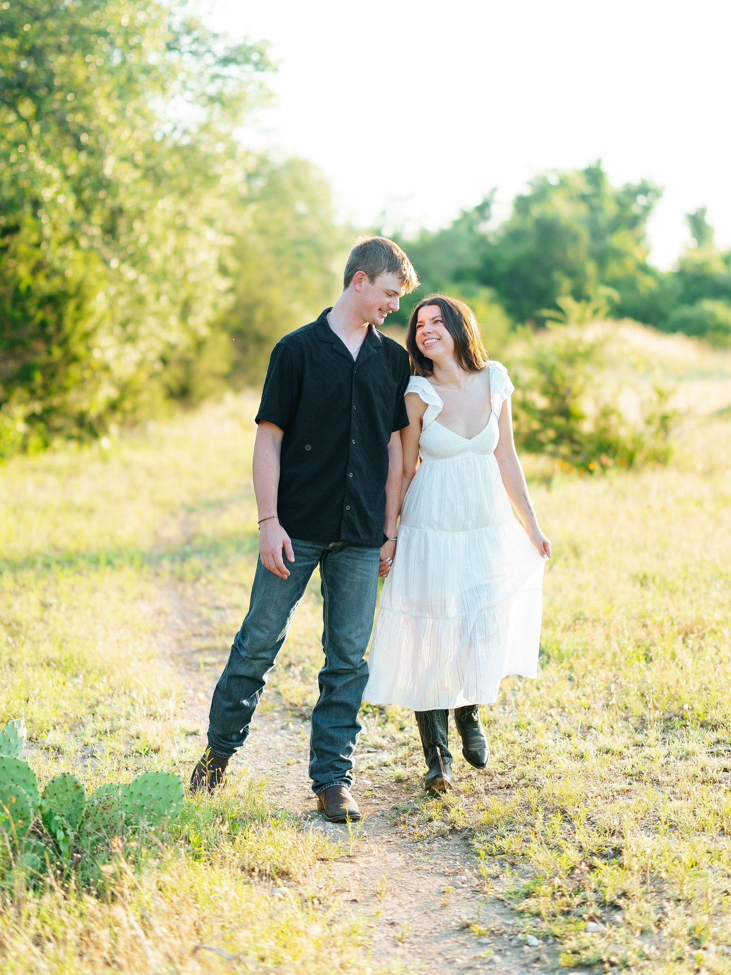 Happy couple walking hand in hand through a sunlit meadow, woman in white dress and man in black shirt.