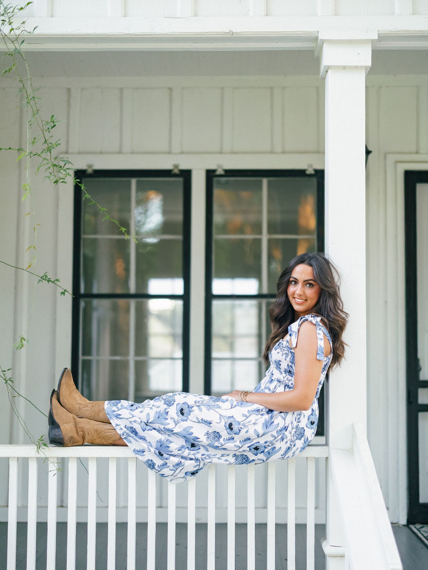 A young woman in a blue floral midi dress and tan boots sits on a white porch railing smiling.