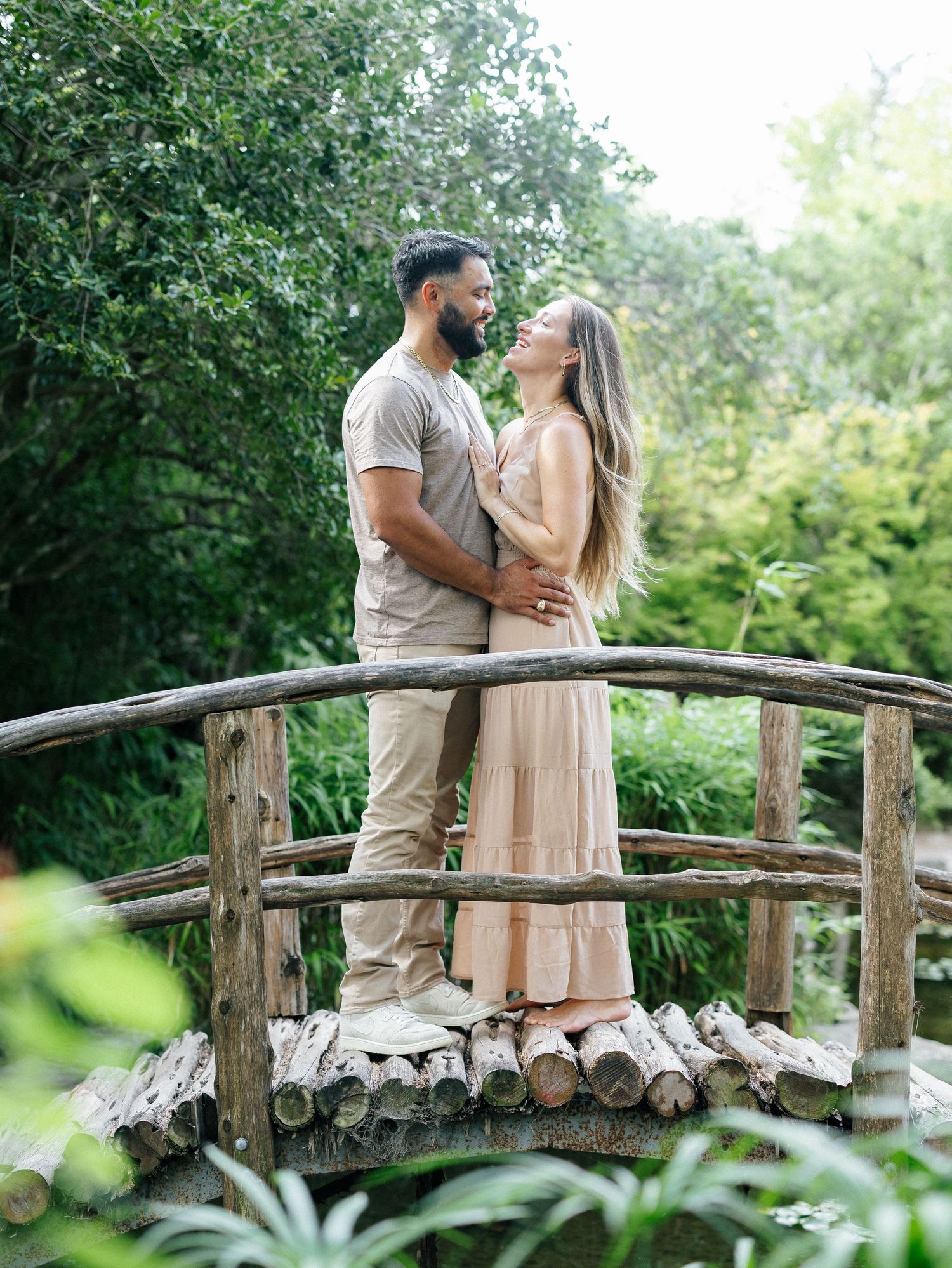 Couple standing close on a rustic wooden bridge surrounded by lush green trees.