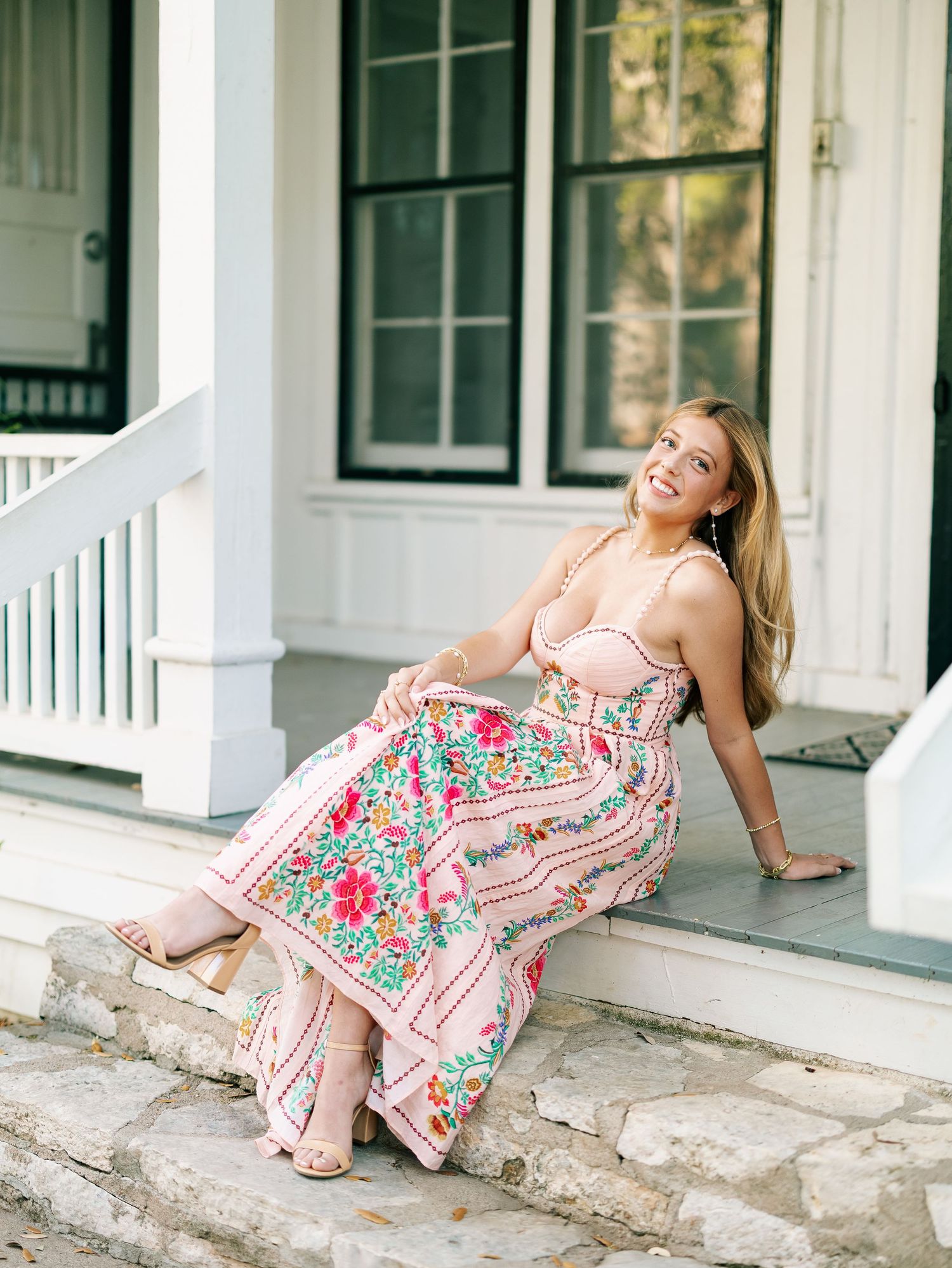 Blonde woman in pink floral maxi dress sitting on stone porch steps smiling.