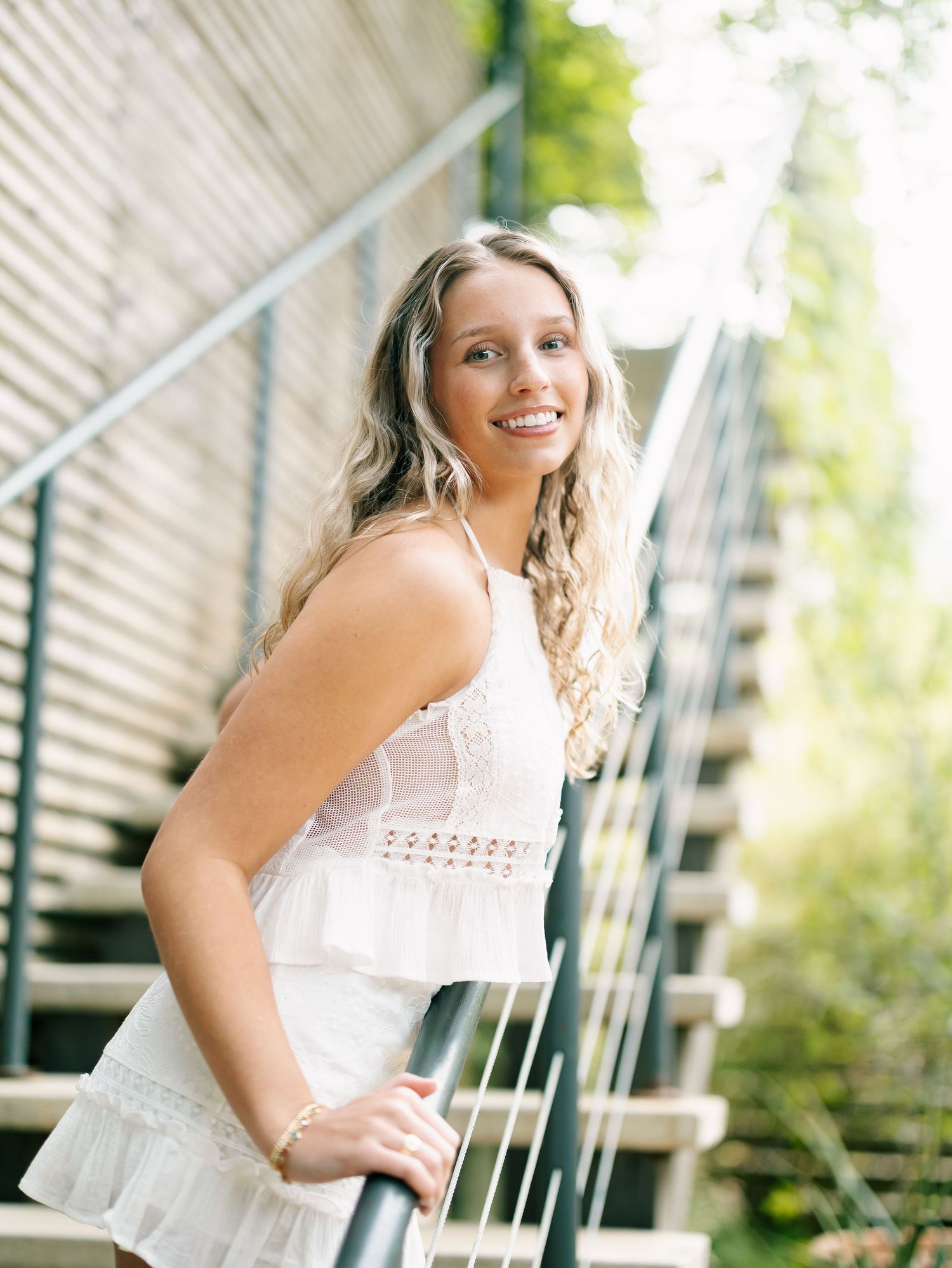 Blonde woman in white lace dress smiling while leaning on outdoor metal staircase railing.