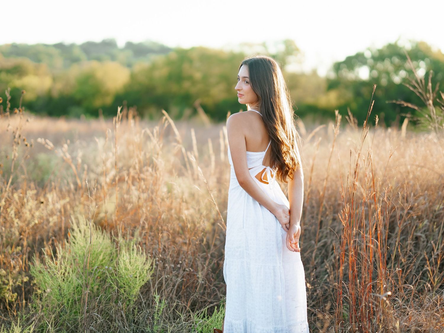 Young woman in white maxi dress standing in golden field at sunset, looking over shoulder.