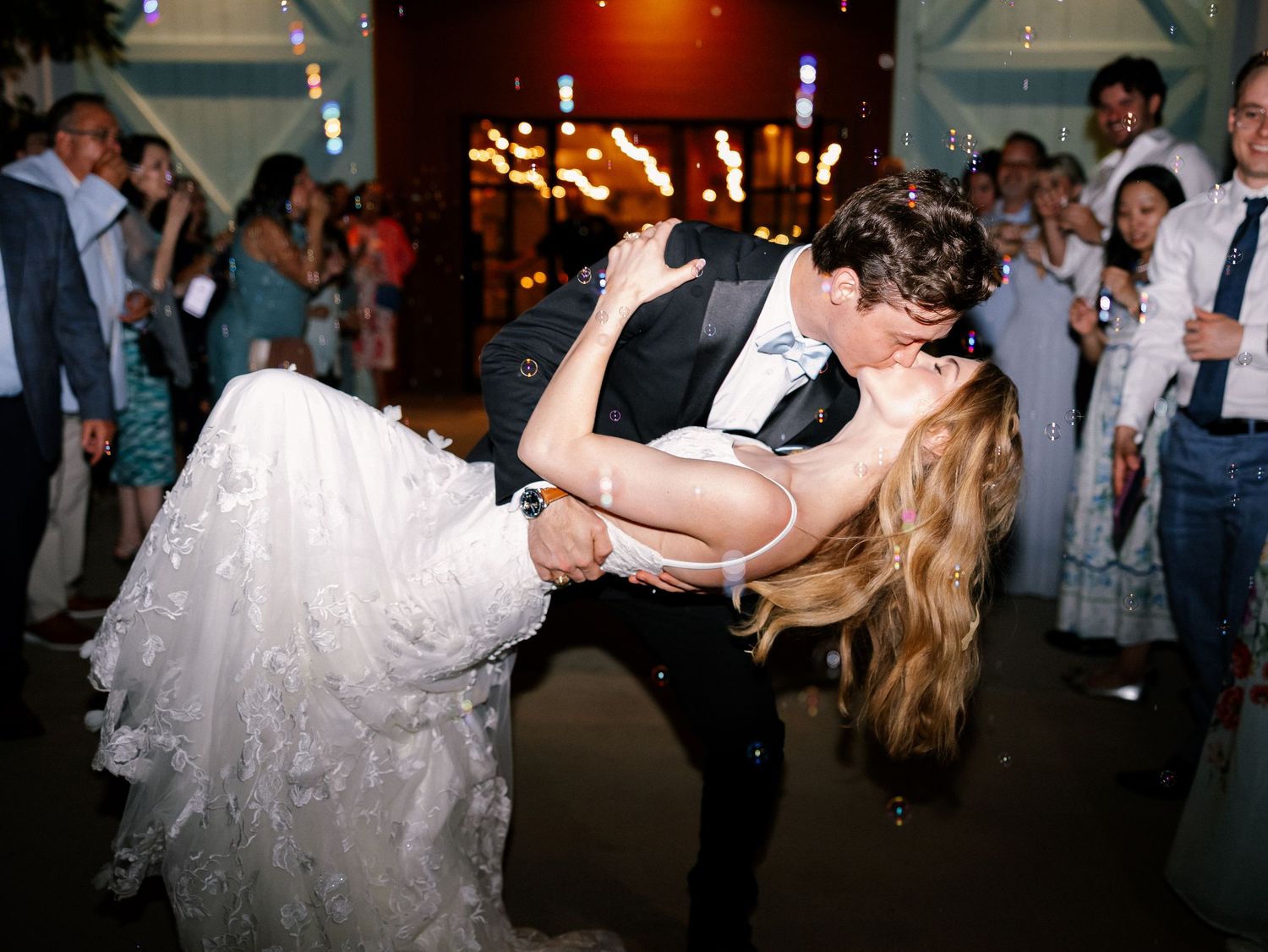 Groom dips bride in wedding dress during lively outdoor reception with sparklers and guests.