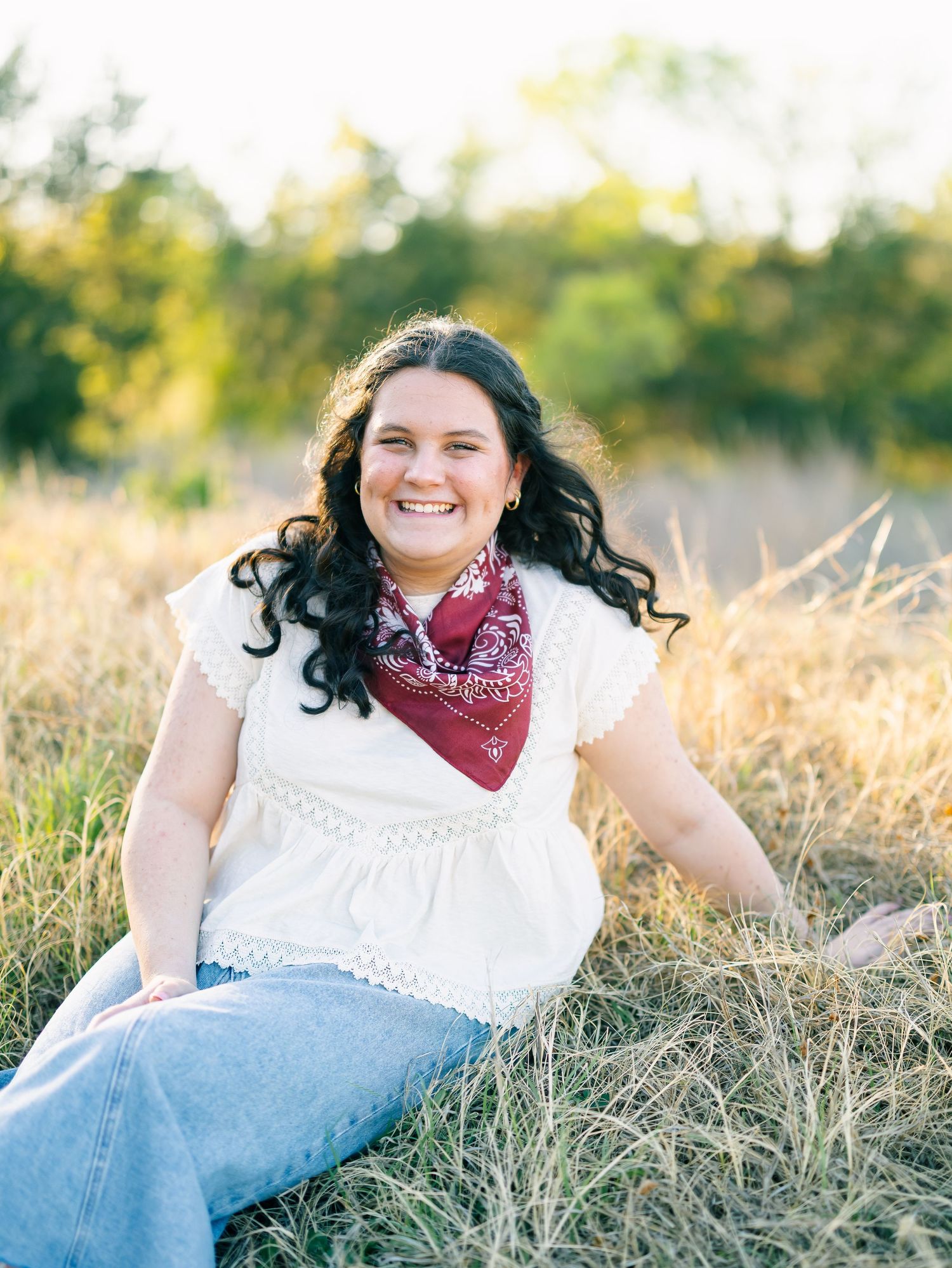 Smiling young woman in white top, red bandana, and jeans sitting in golden field with trees in background.