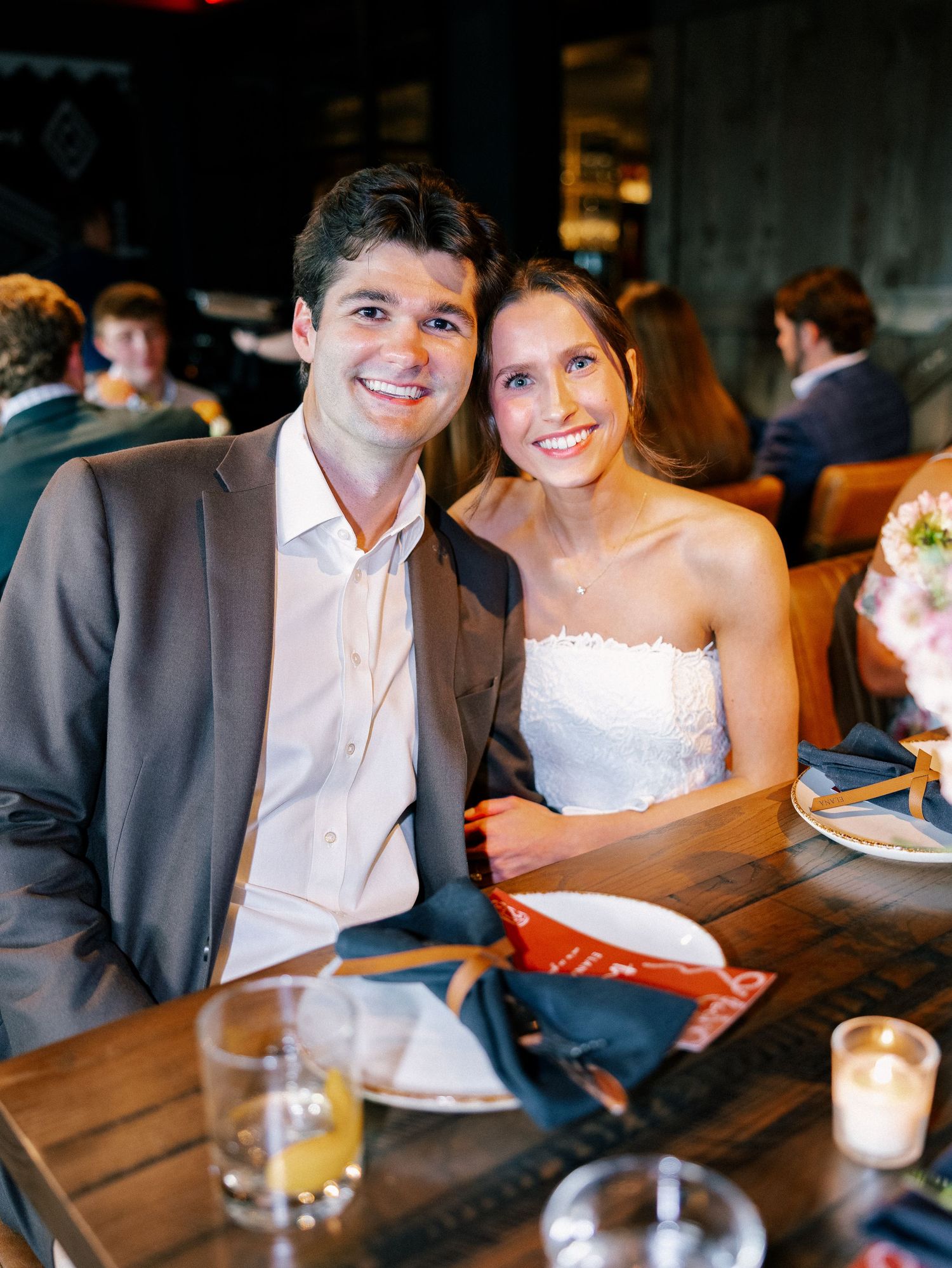 Smiling bride in strapless gown and groom in gray suit seated at wedding reception table.