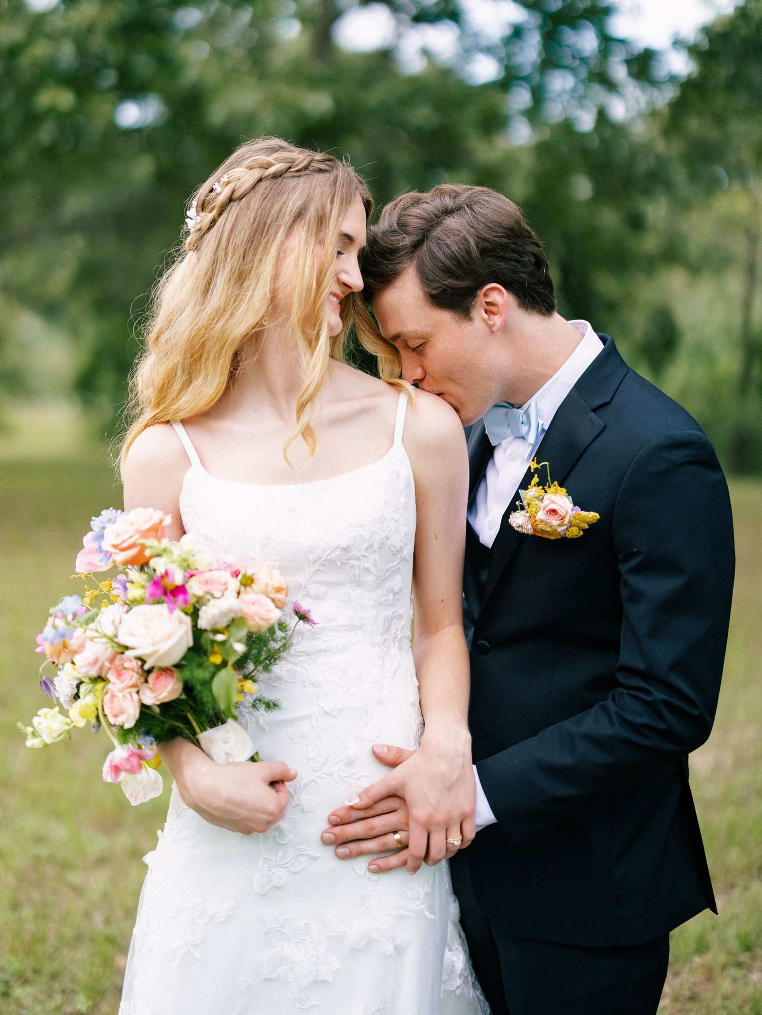 Bride and groom share an intimate moment outdoors, bride holding colorful bouquet in white lace gown, groom in navy suit.