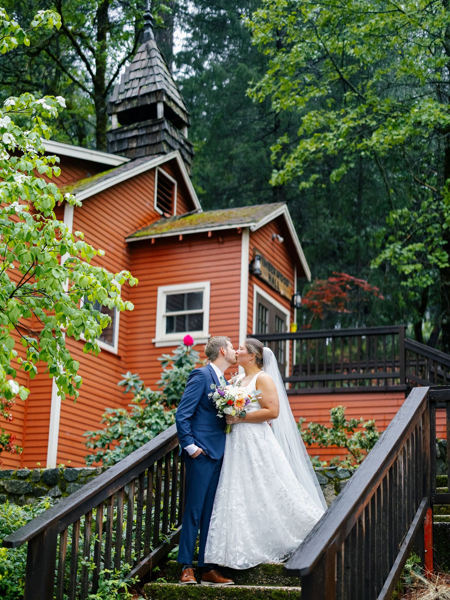 Bride and groom kissing on wooden bridge steps before a charming red chapel surrounded by lush green trees.