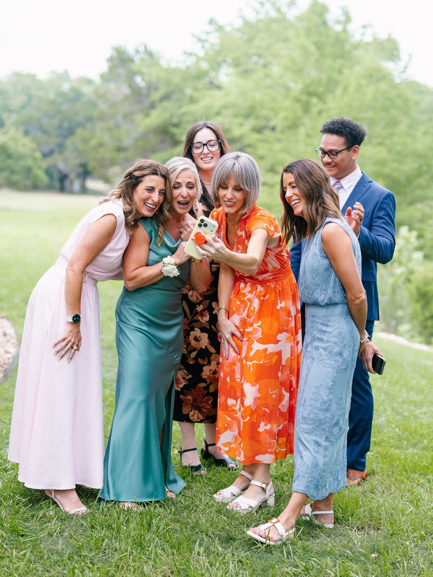 Group of elegantly dressed friends laughing and looking at a phone outdoors at a wedding celebration.