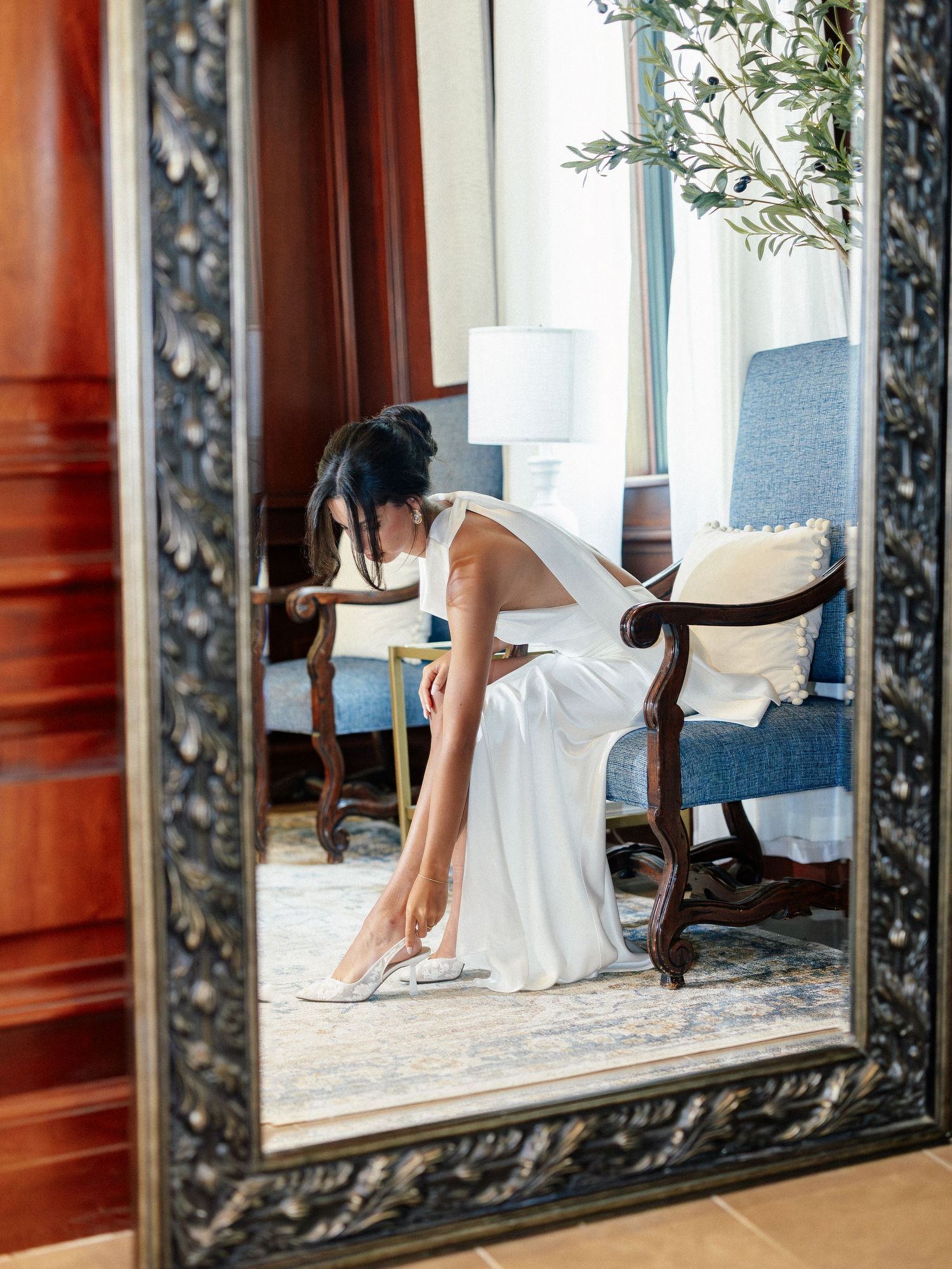 Bride in white gown putting on heels, reflected in ornate mirror in elegant room.