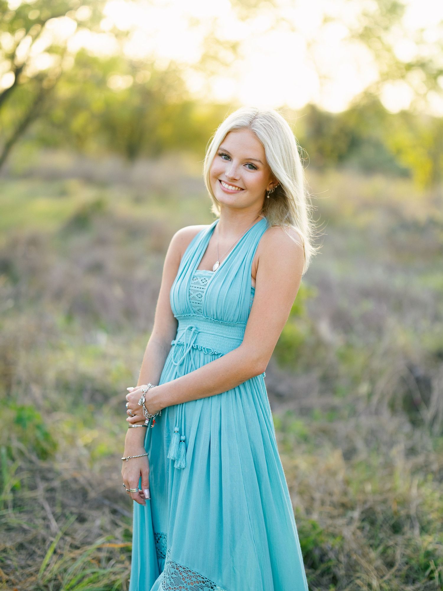 Smiling blonde woman in turquoise halter maxi dress posing in golden hour sunlit field.