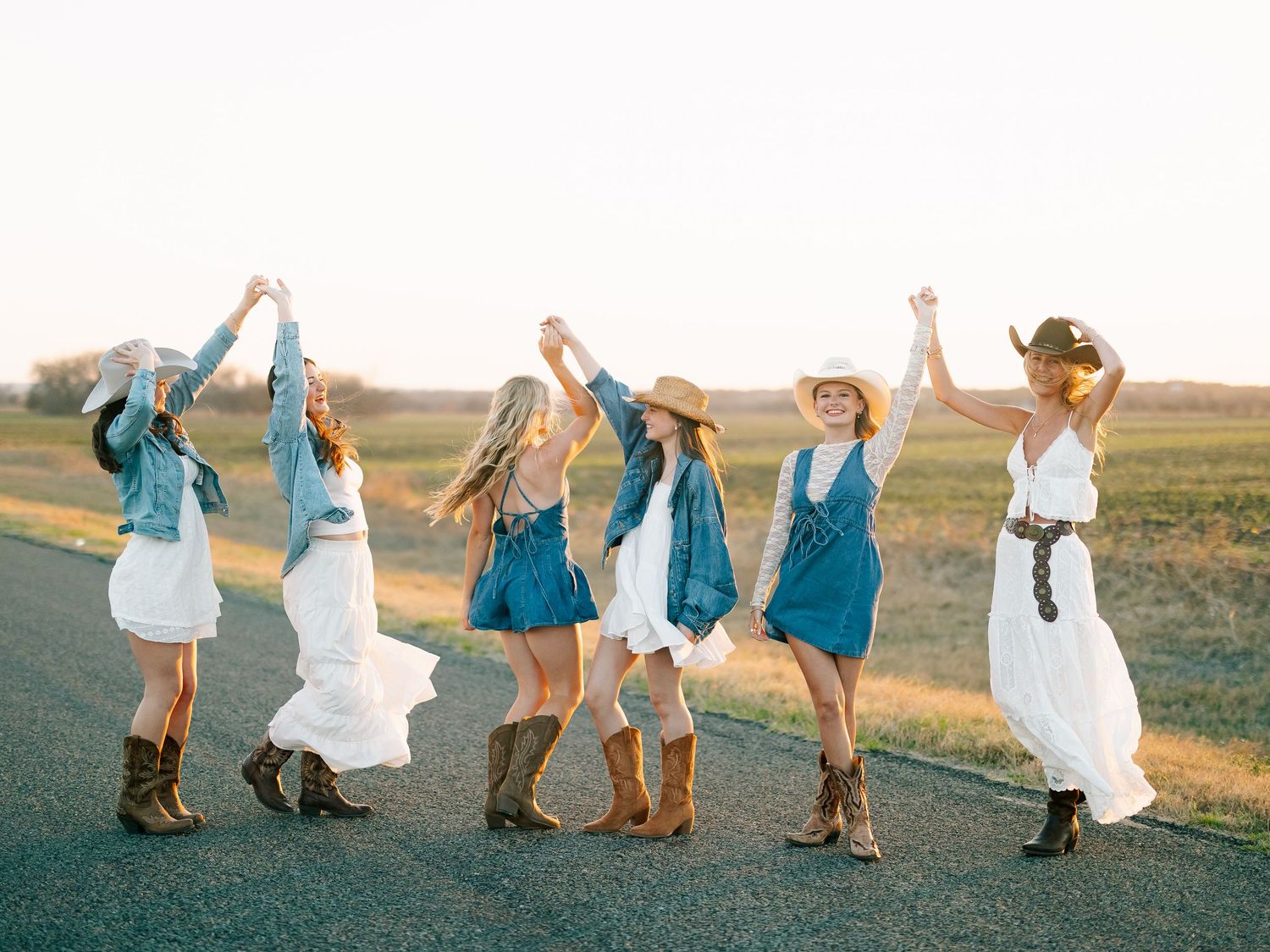 Six women in cowboy boots and hats dance joyfully on a road at sunset, wearing white dresses and denim outfits.