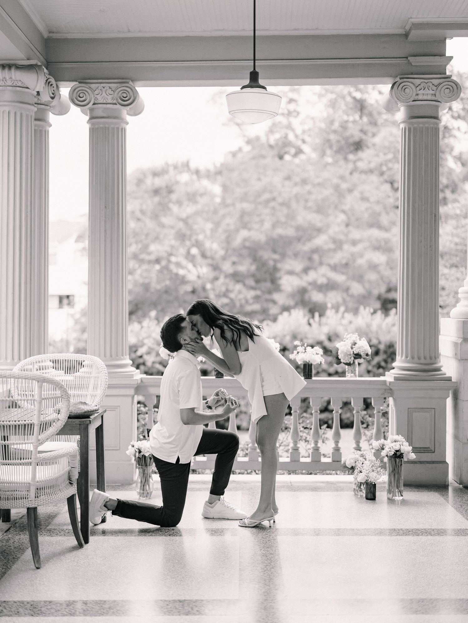 Black and white photo of a man on one knee proposing to his partner on a grand columned porch.
