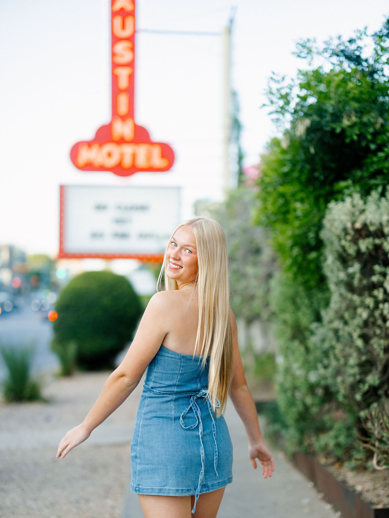 Young blonde woman in denim mini dress smiling in front of Austin Motel neon sign on South Congress Avenue.