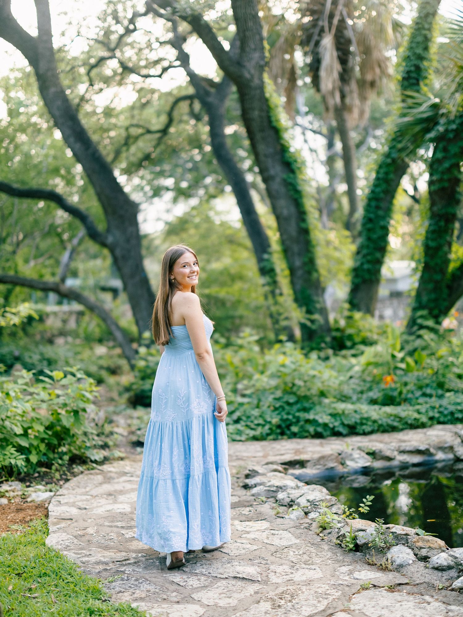 Brunette woman in light blue maxi dress smiling on a stone path beside a garden pond.