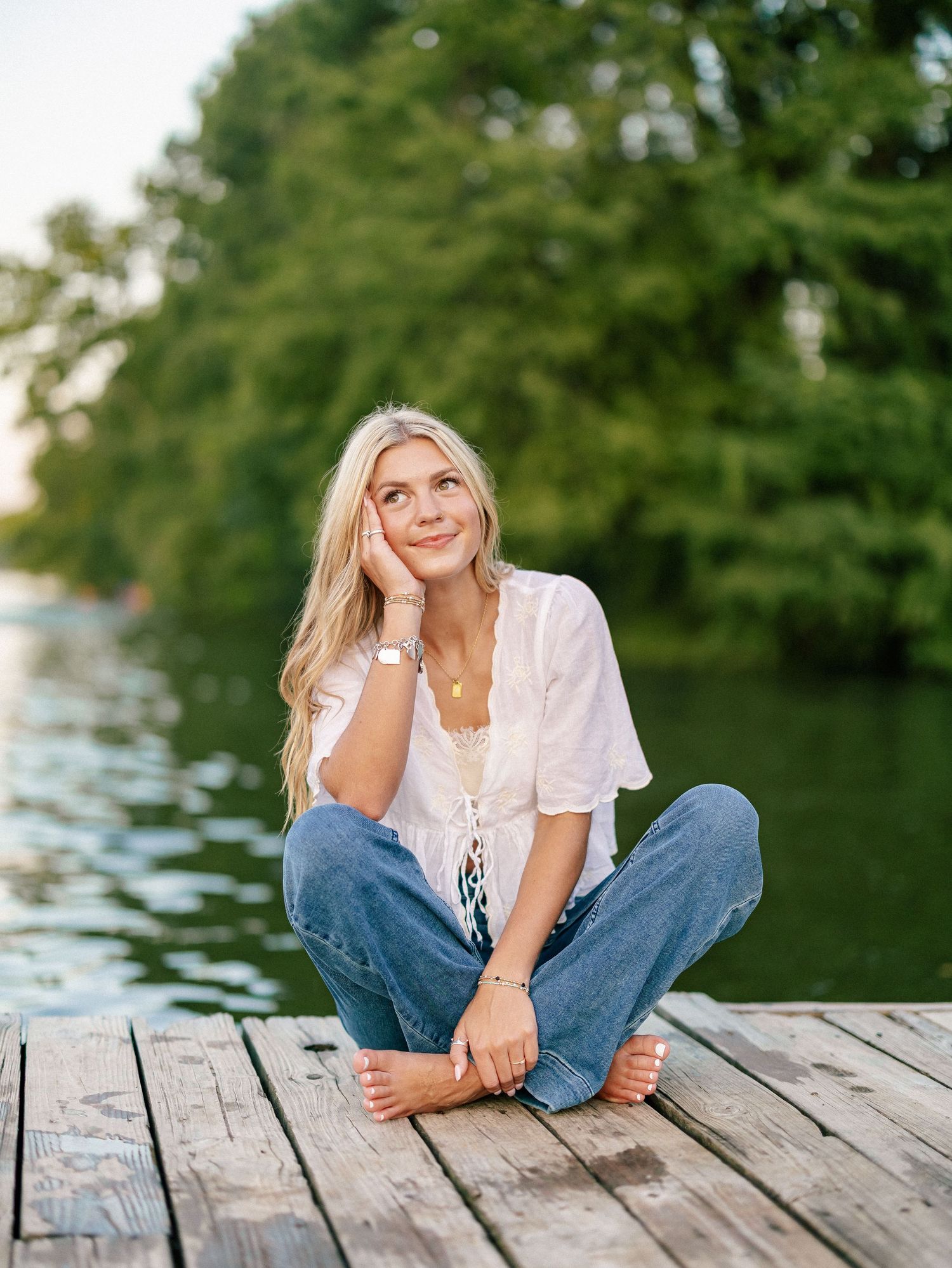 Blonde woman sitting cross-legged on a wooden dock by a lake, wearing jeans and white blouse, smiling dreamily.