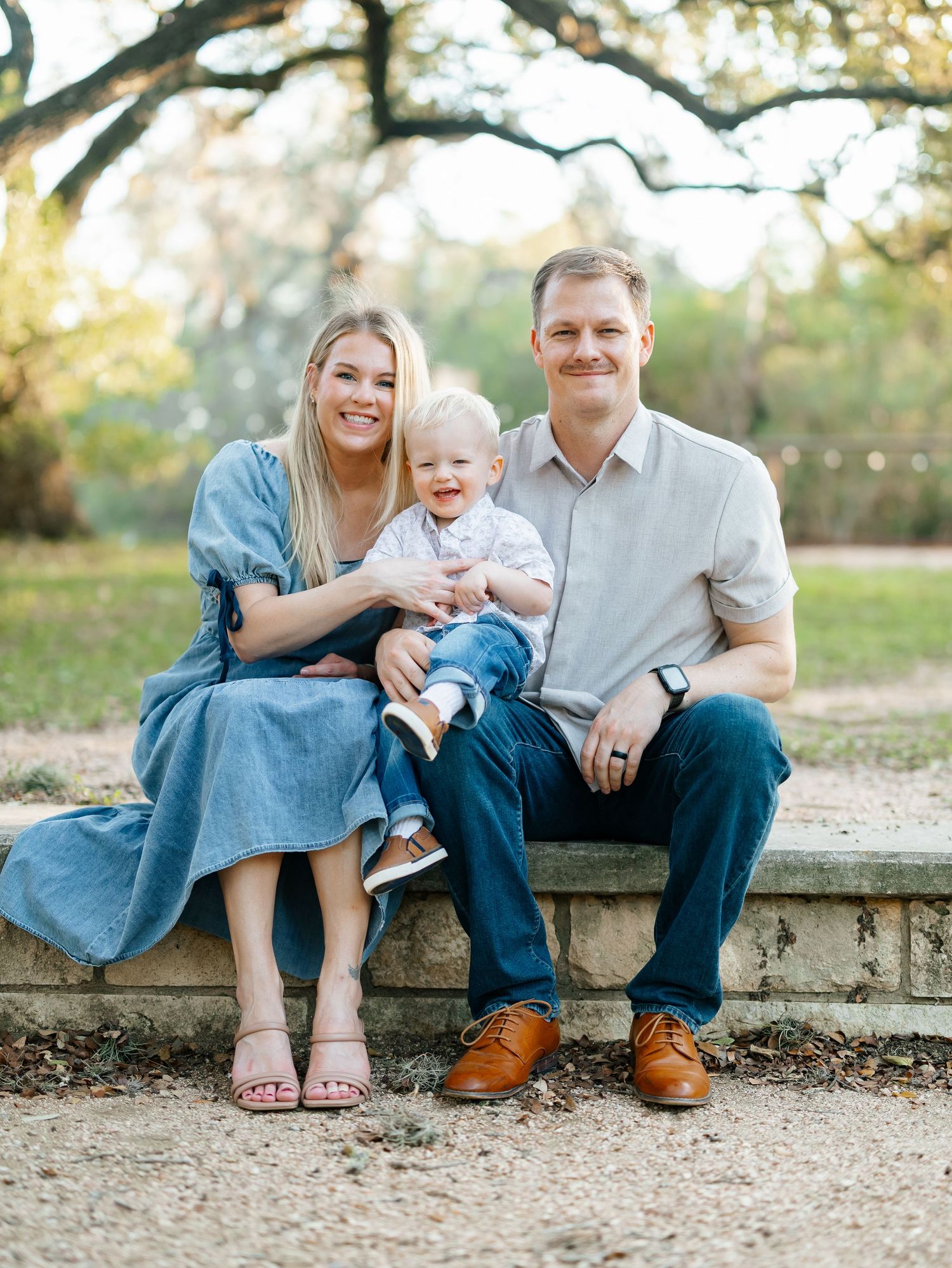 Smiling family with baby boy seated on stone bench in a sunlit park setting.