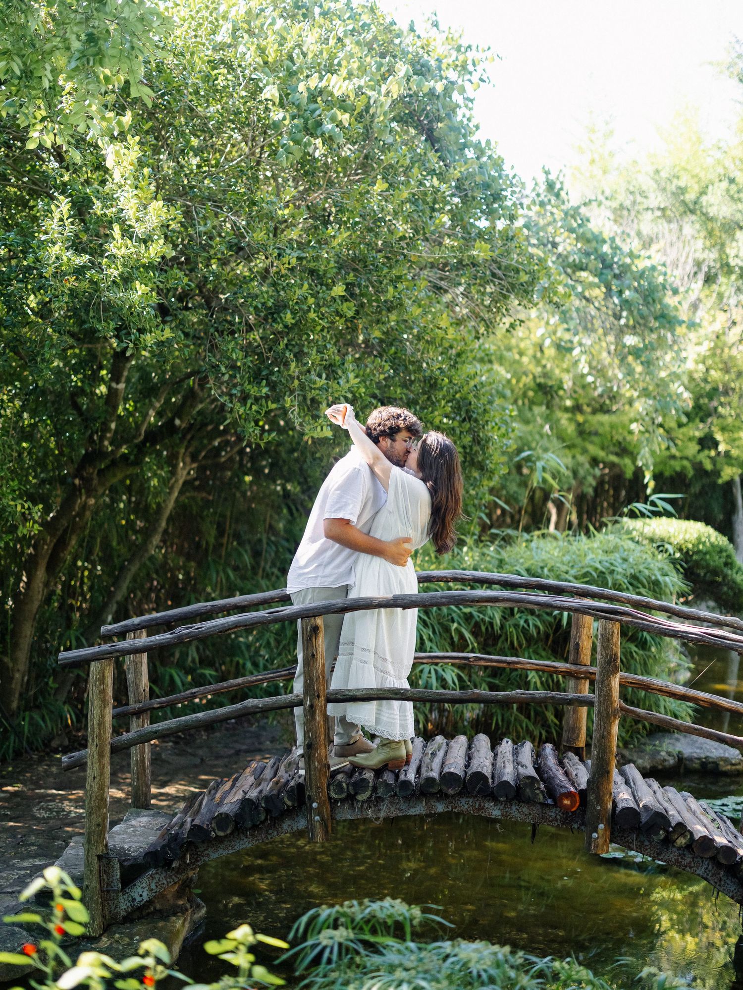 Couple sharing a romantic kiss on a rustic wooden bridge surrounded by lush green trees in a garden.