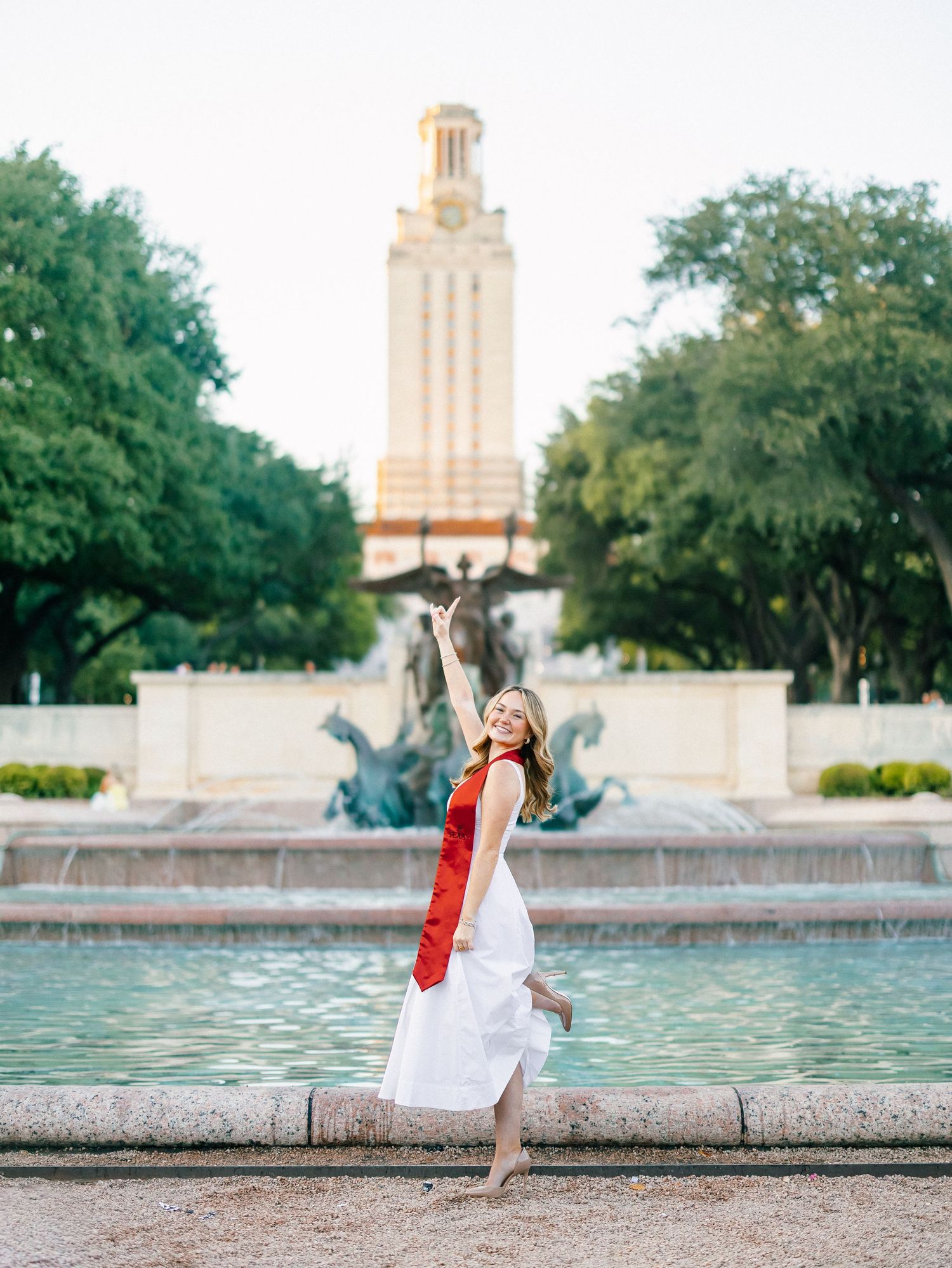 Graduate in white dress and red sash poses joyfully by UT Austin fountain with the iconic tower backdrop.
