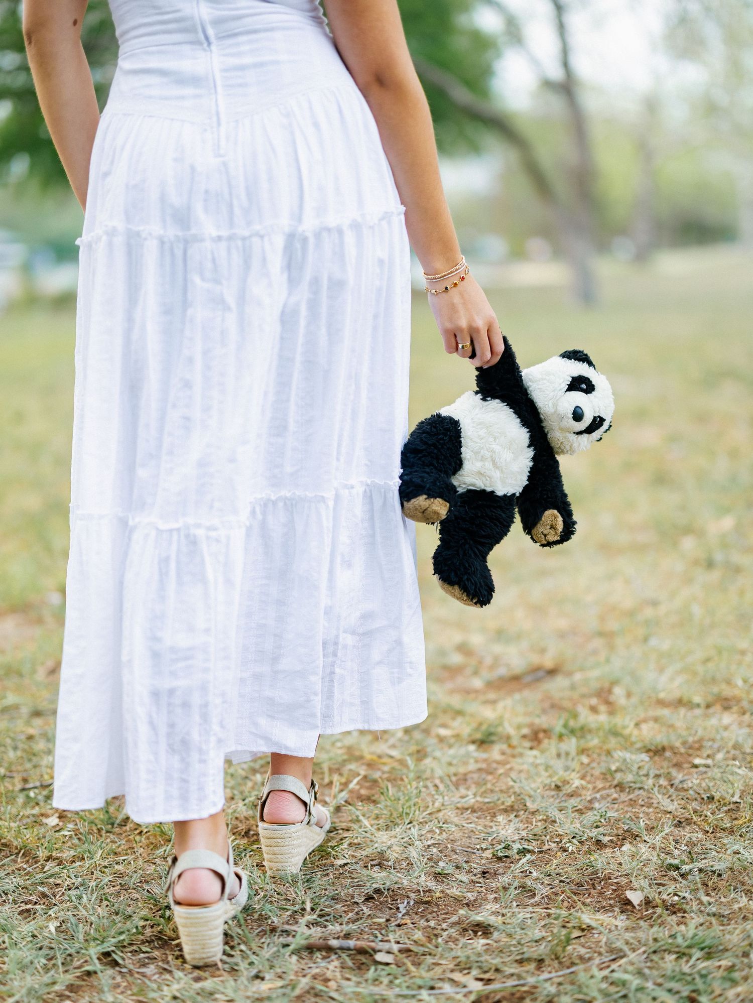 Woman in white tiered maxi skirt holding a black and white panda stuffed animal toy while standing on grass.