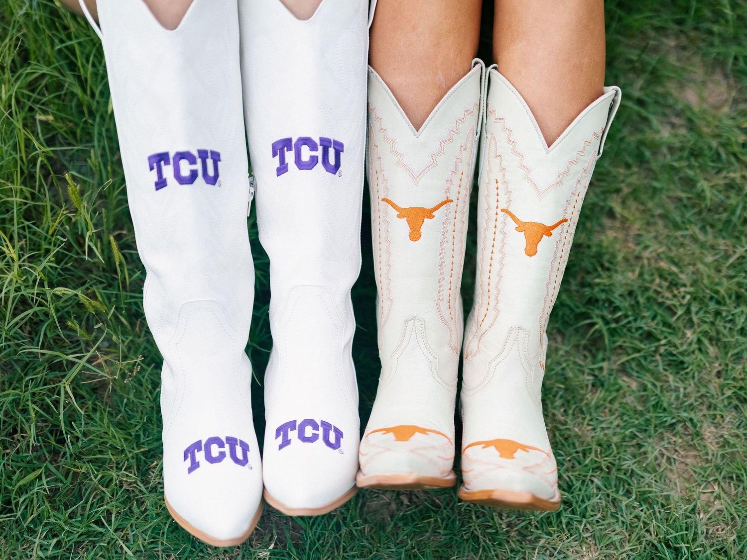 Two pairs of white cowboy boots featuring TCU purple logos and University of Texas longhorn emblems on green grass.