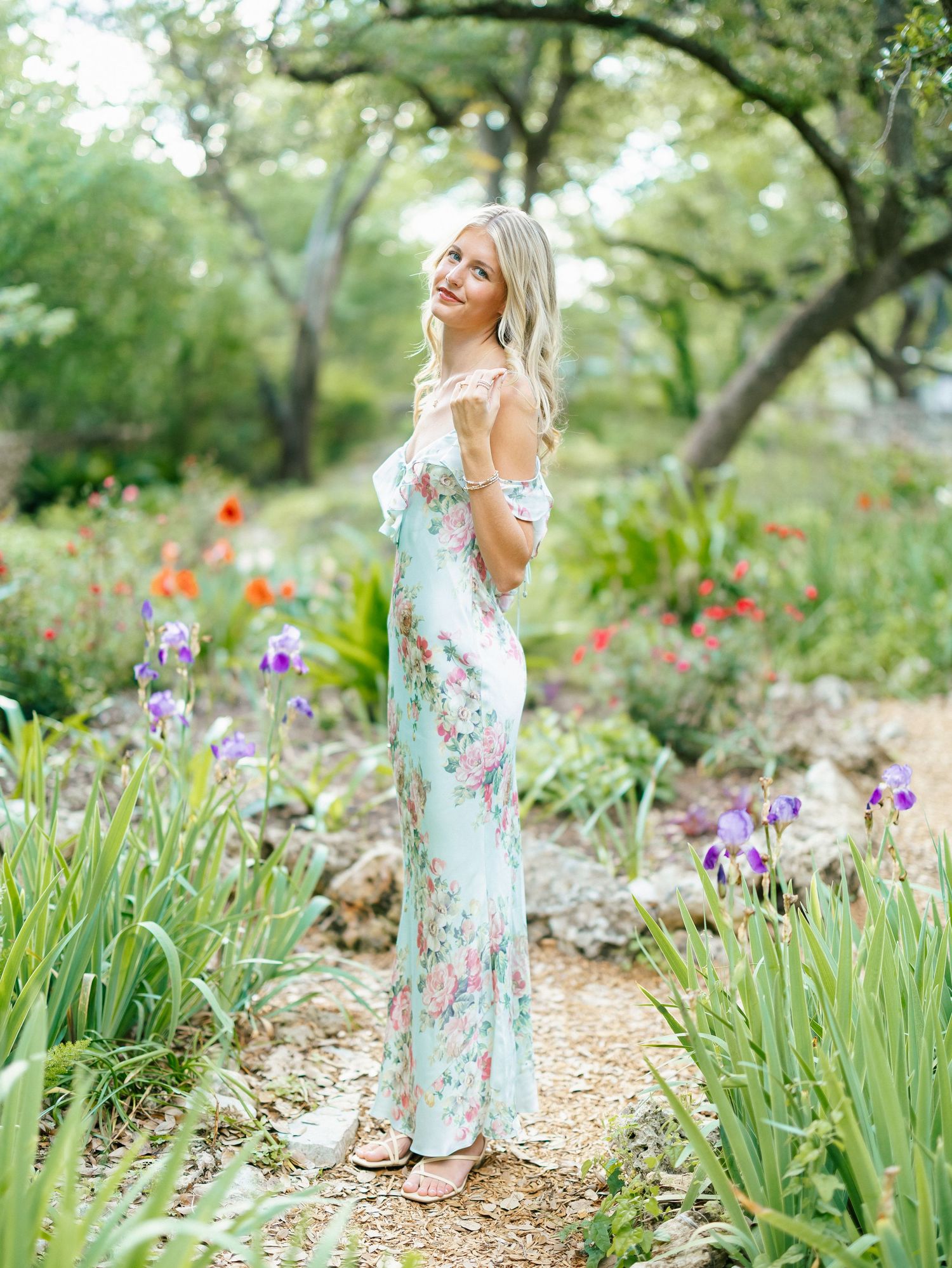 Blonde woman in a floral mint maxi dress standing on a garden path surrounded by colorful iris flowers and lush greenery.