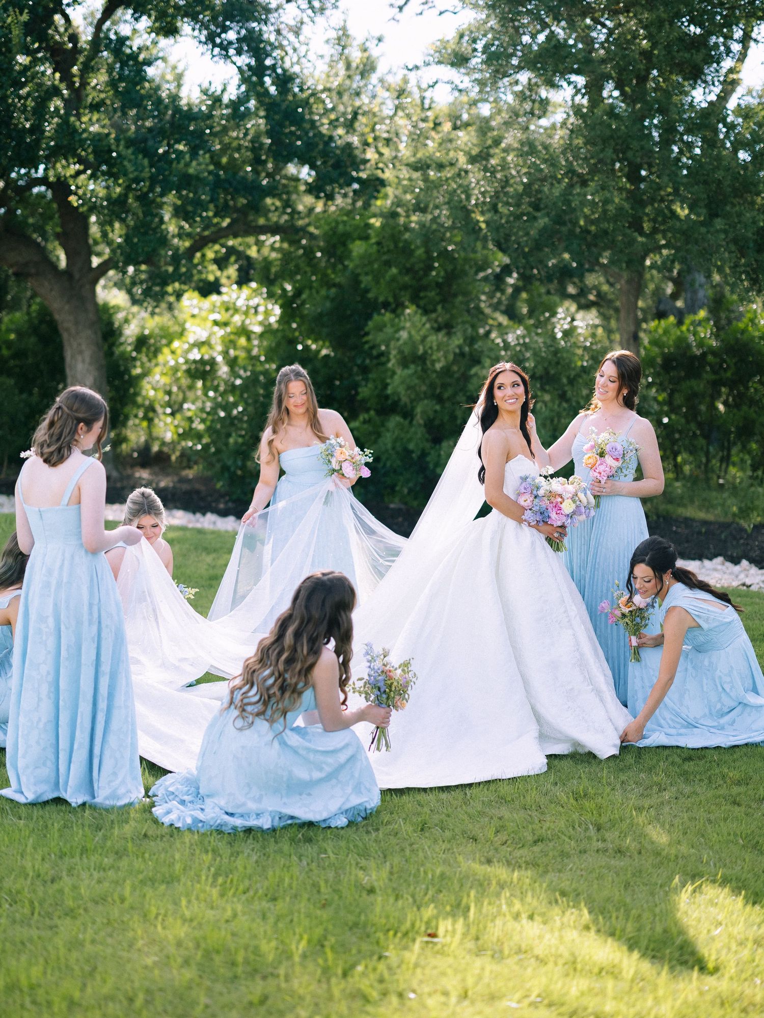 Bride in white gown with bridesmaids in light blue dresses arranging her train on green lawn outdoors.