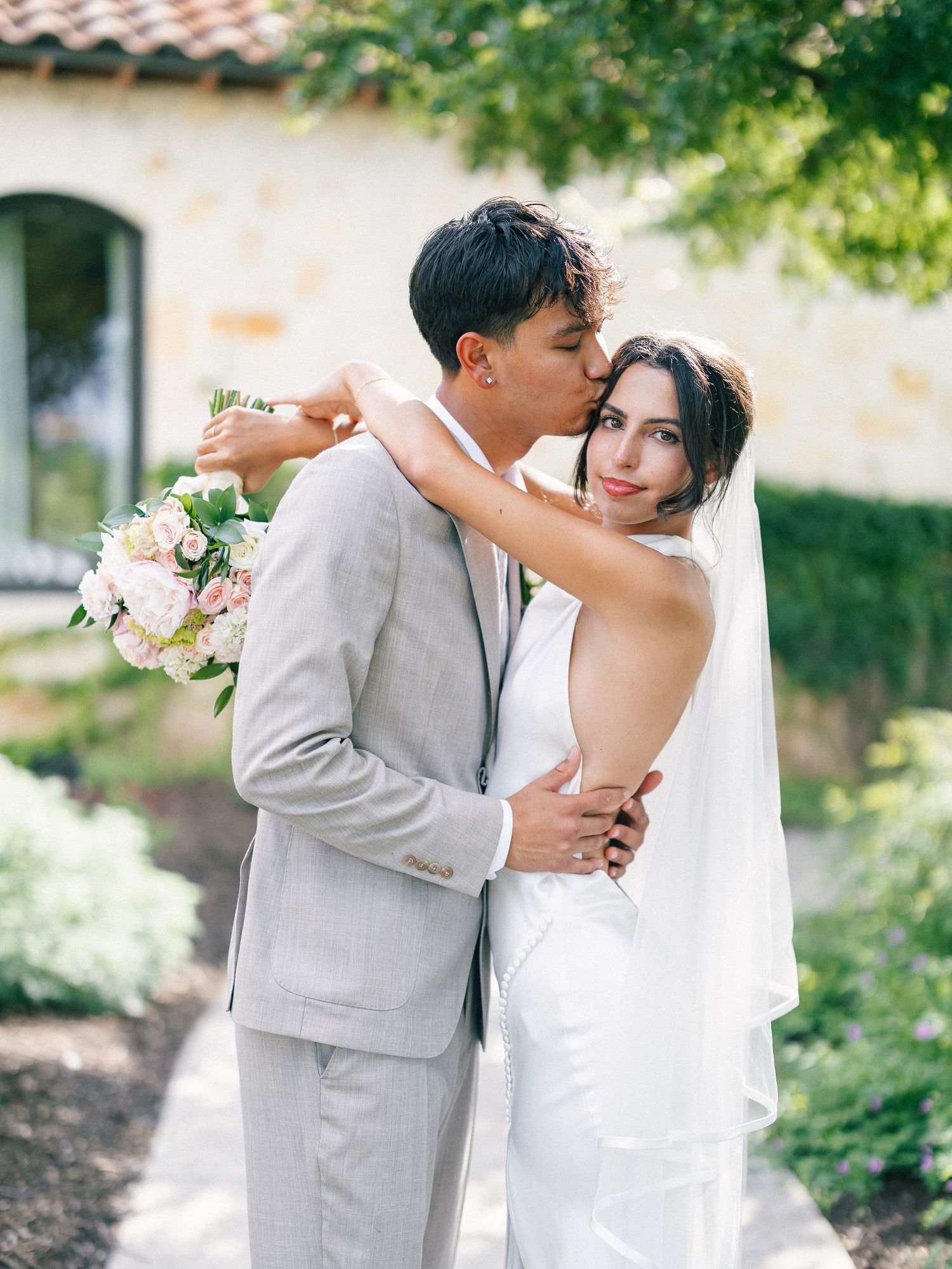 A groom in a gray suit kisses his bride wearing a white gown and veil while she holds a bouquet outdoors.